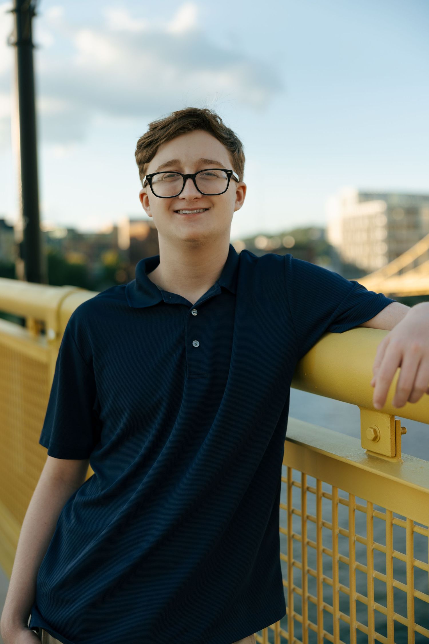 Teen boy with glasses smiling on yellow bridge railing outdoors.