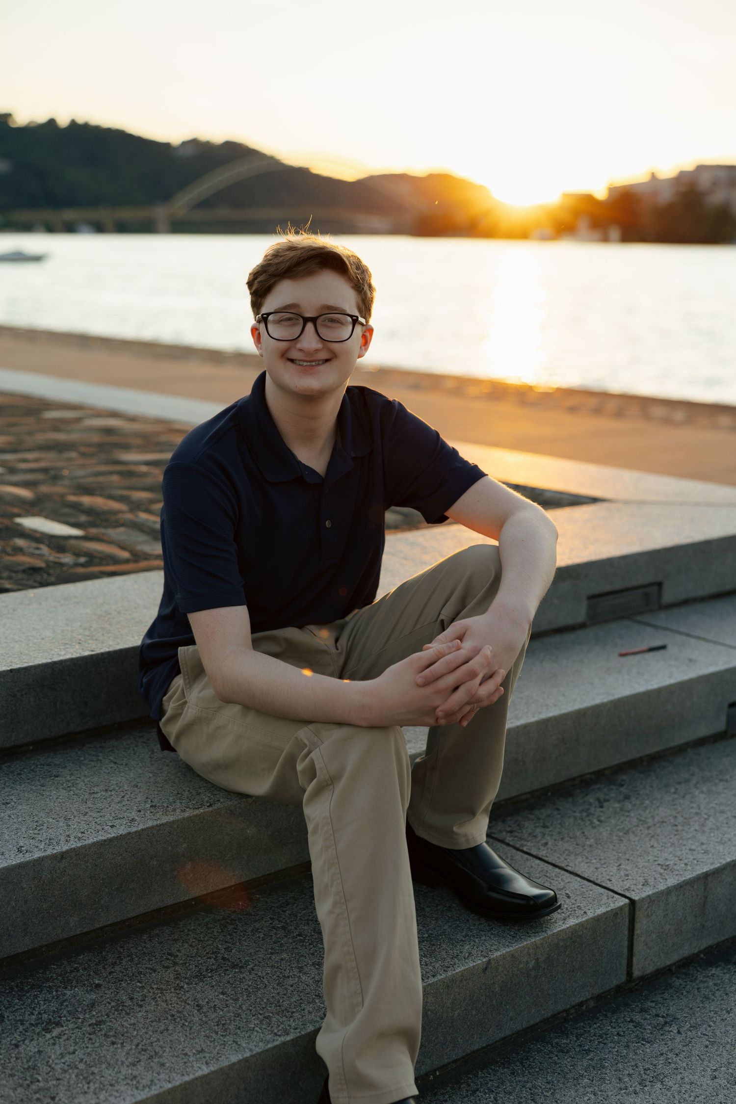 Smiling teenage boy sitting on stone steps by a river at sunset with golden light behind him.