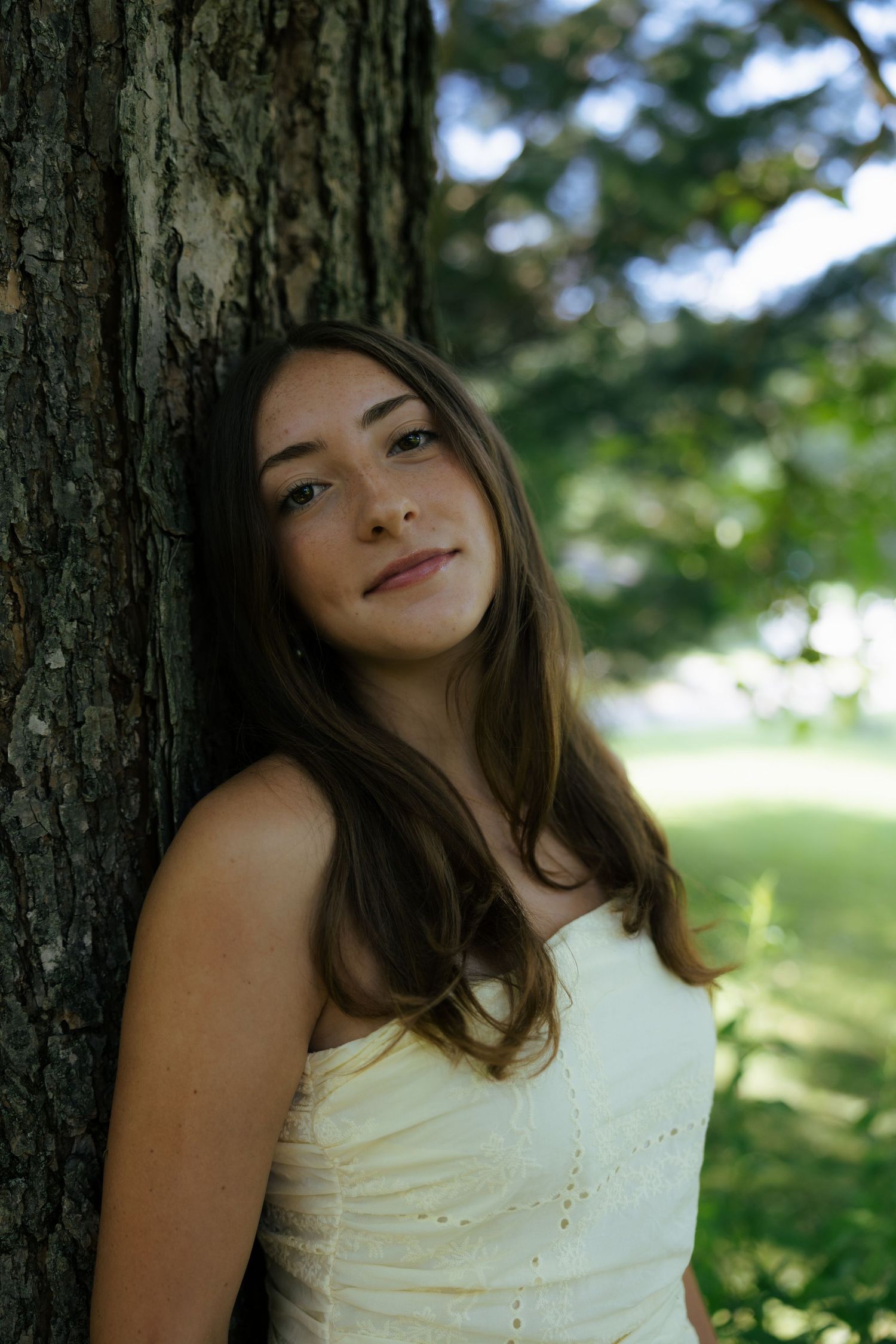 Young woman in white dress leaning against tree in lush green park, smiling softly at camera.