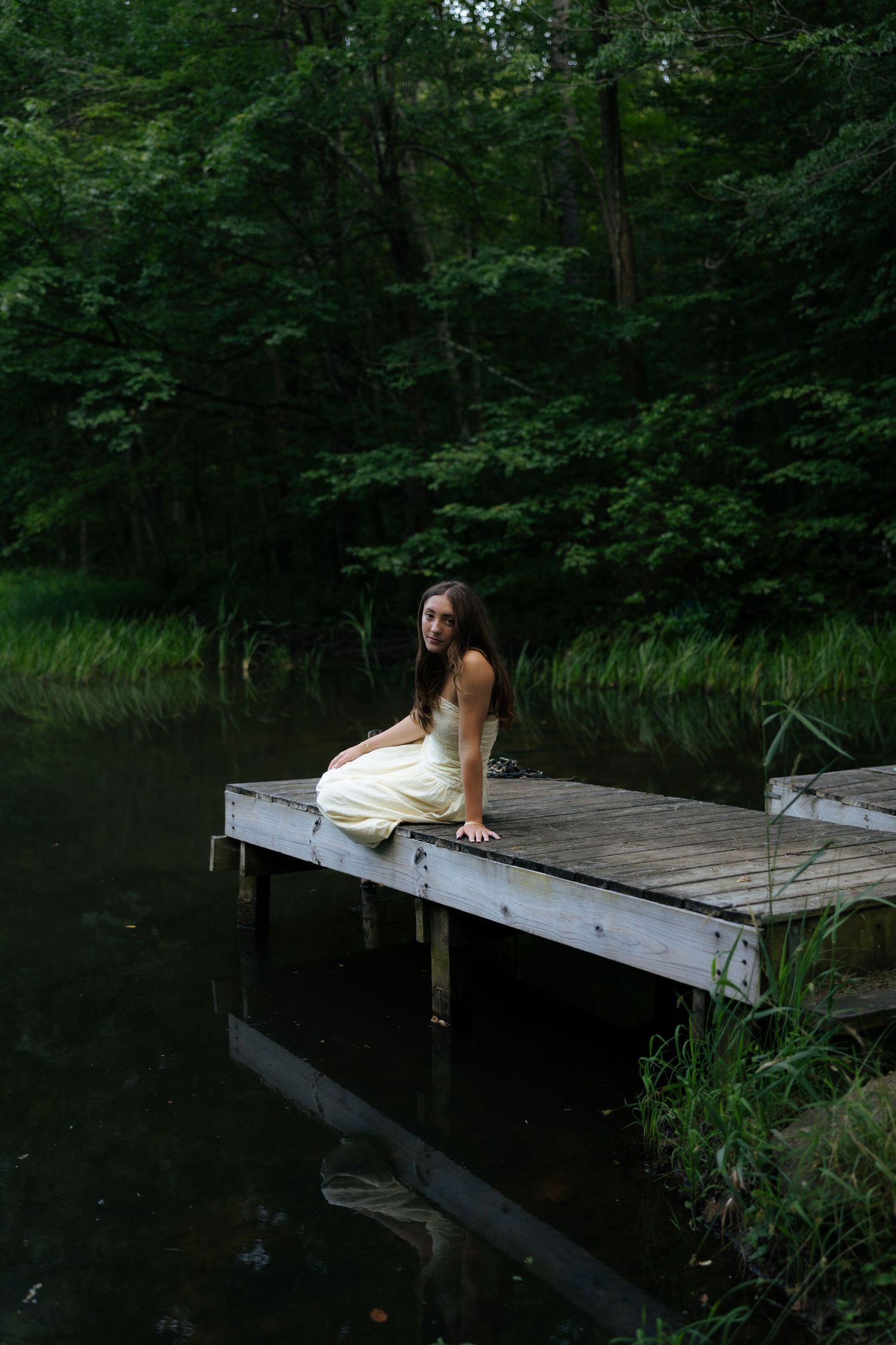 Young woman in white dress sitting on edge of wooden dock over dark lake in forest.