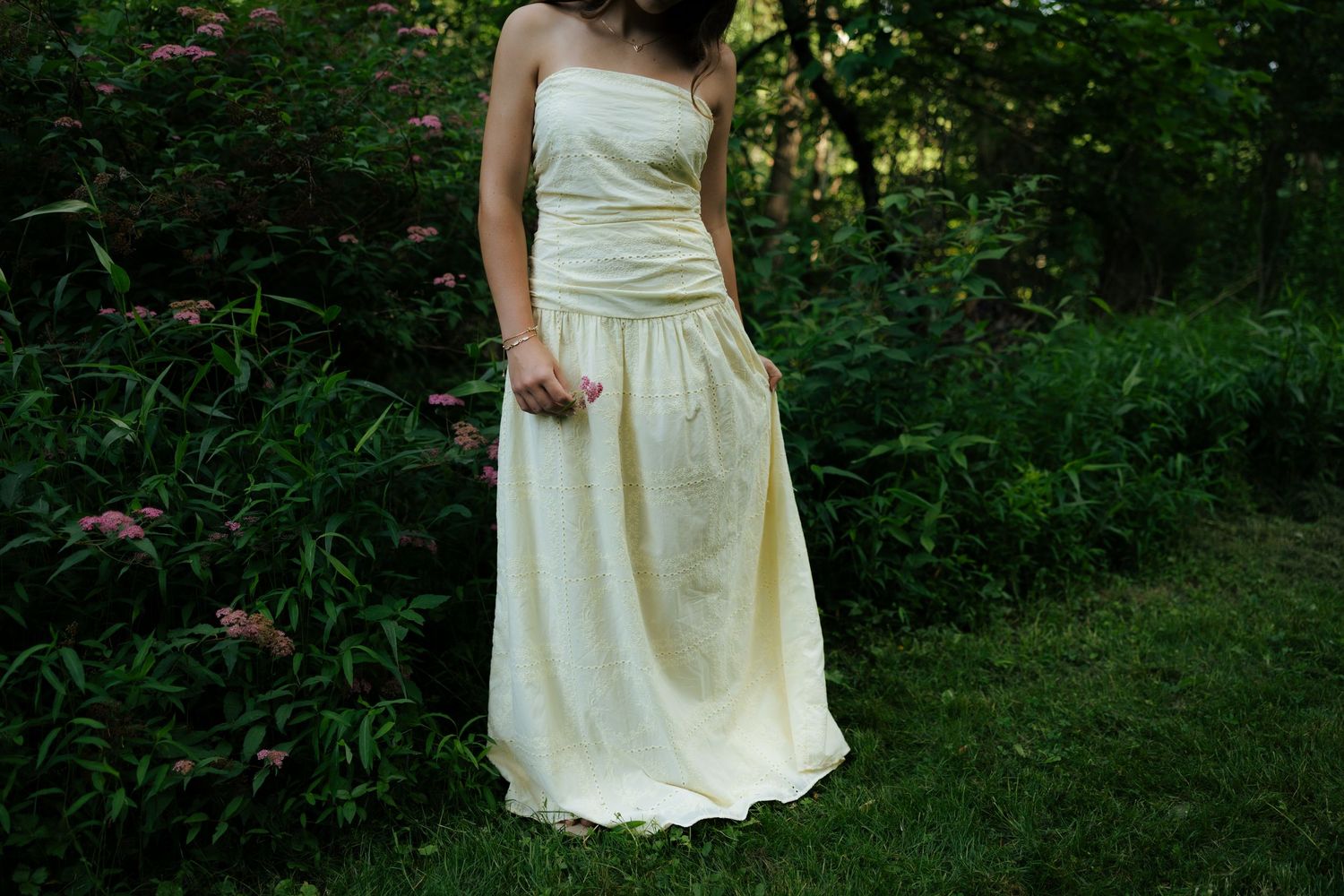 Cream strapless maxi dress worn by woman standing in garden with pink flowering shrubs.