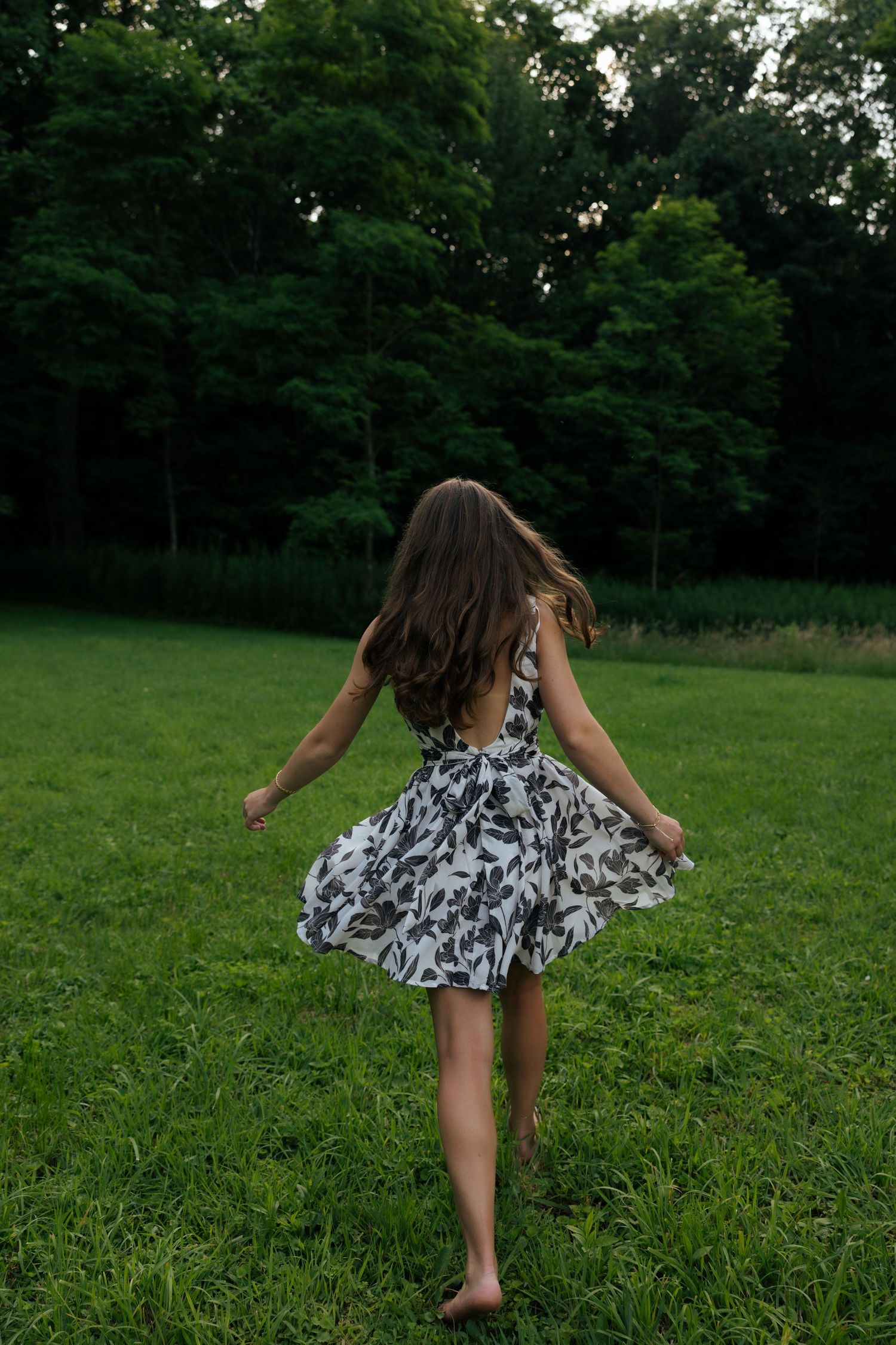 Young woman in black and white floral dress twirling barefoot on lush green grass in a park.