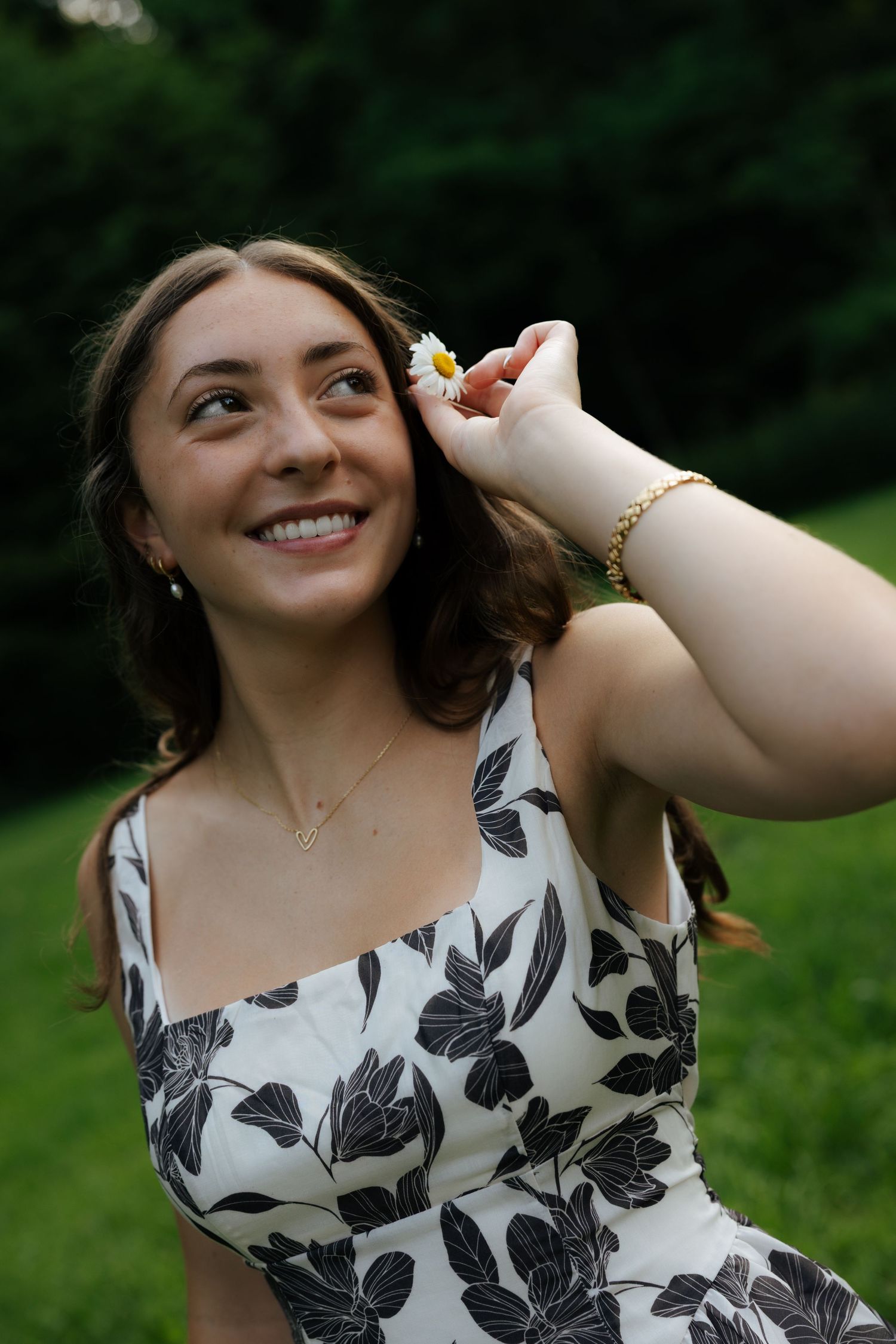 Smiling young woman holding daisy flower near ear in floral dress outdoors.