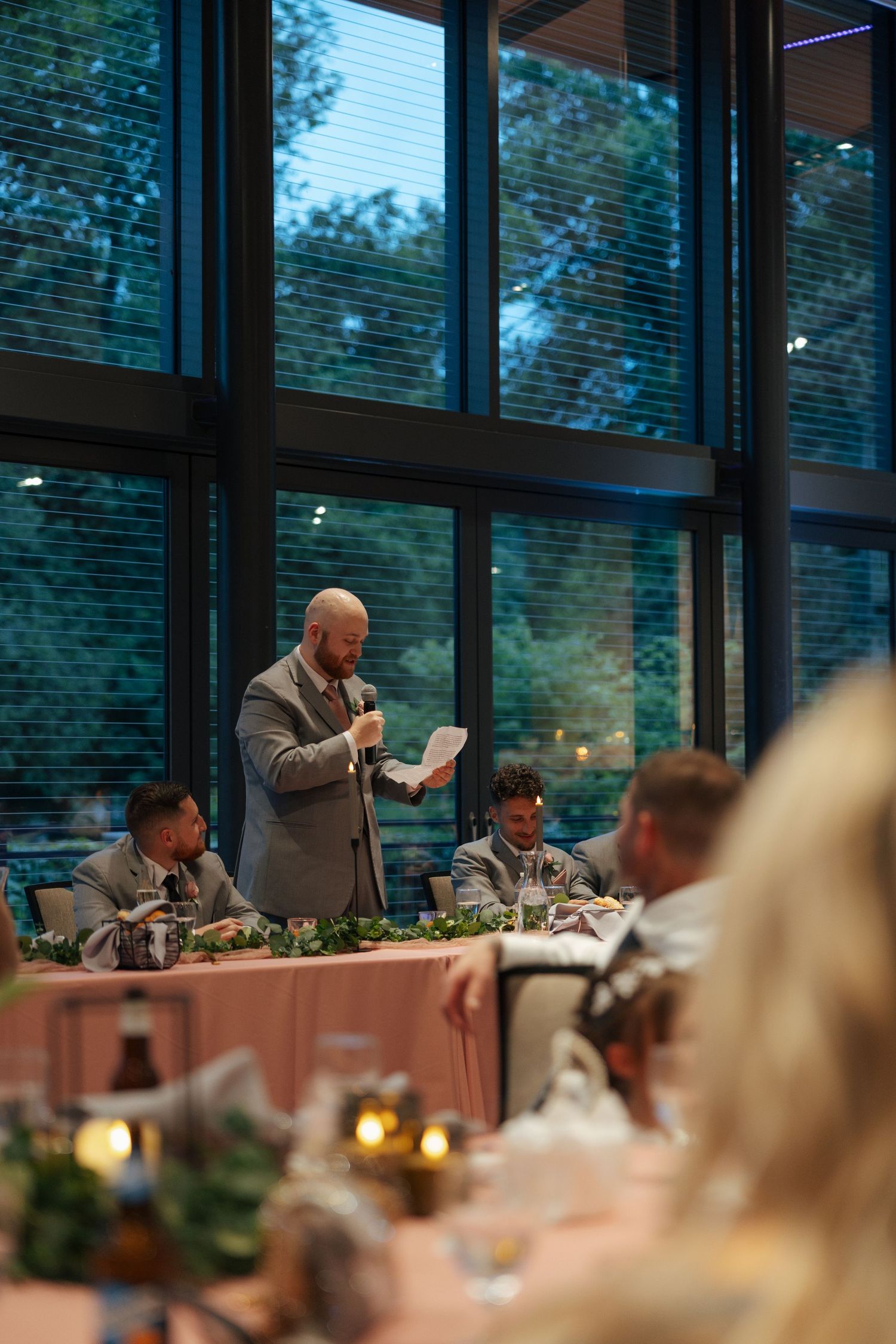 A bearded groomsman in a gray suit delivers a heartfelt wedding speech at an elegant evening reception.