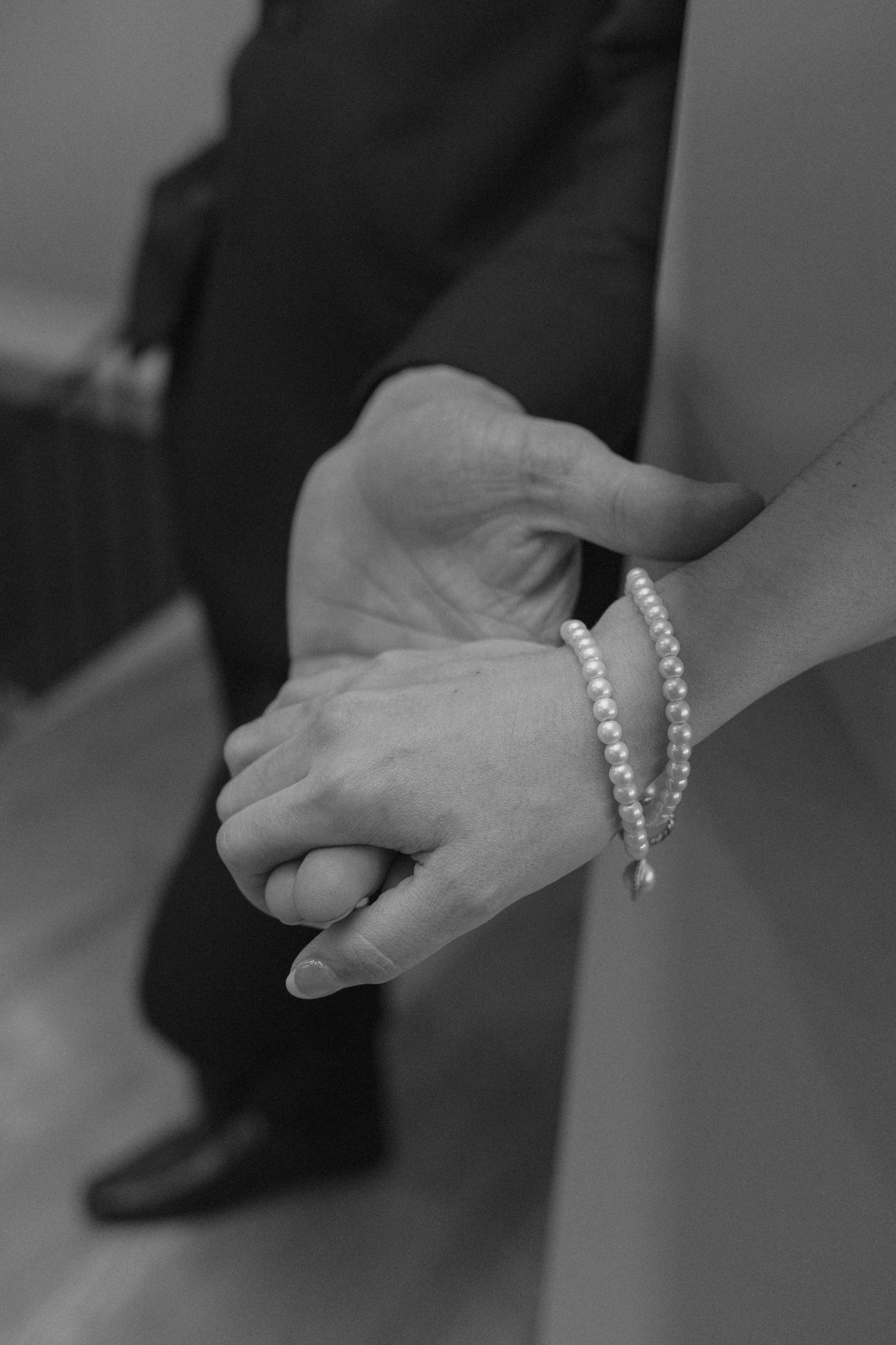Close-up black and white photo of bride and groom holding hands, bride wearing a delicate pearl bracelet.