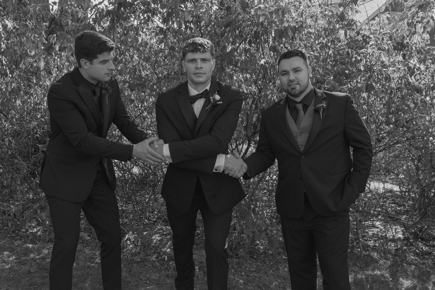 Three men in black tuxedos shaking hands outdoors in a black and white formal wedding photo.