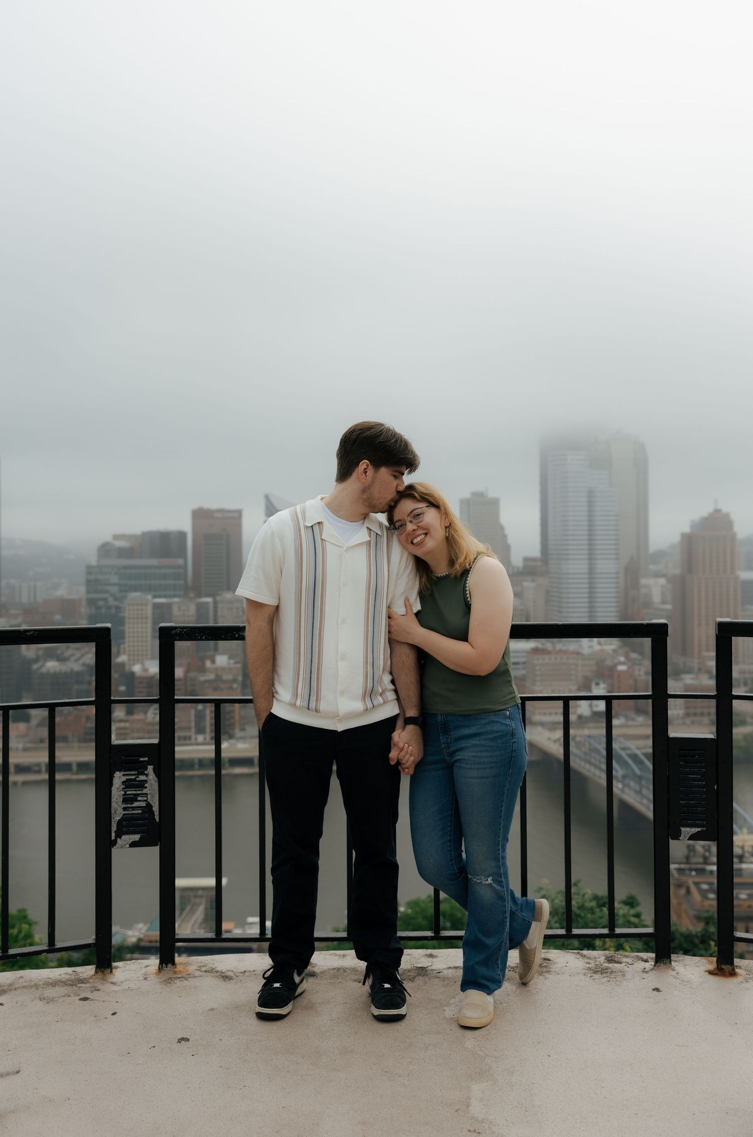 Happy couple embracing on a rooftop overlook with Pittsburgh city skyline in the misty background.