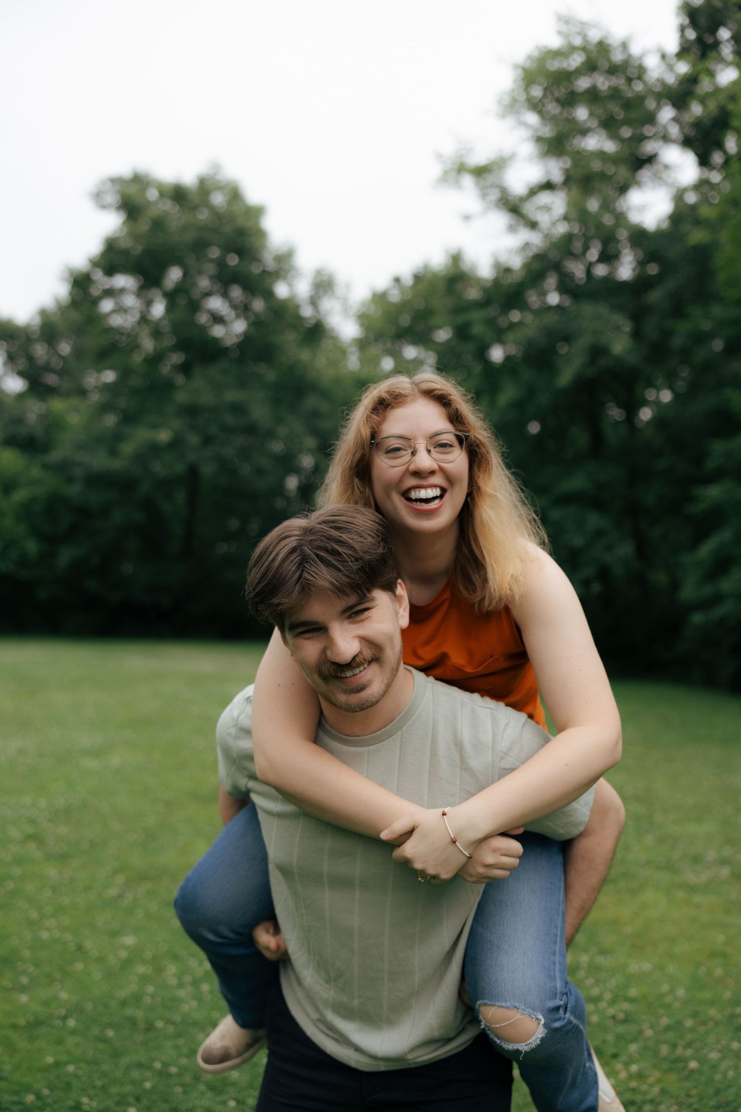 Laughing woman with glasses rides piggyback on smiling man with mustache in a lush green park setting.