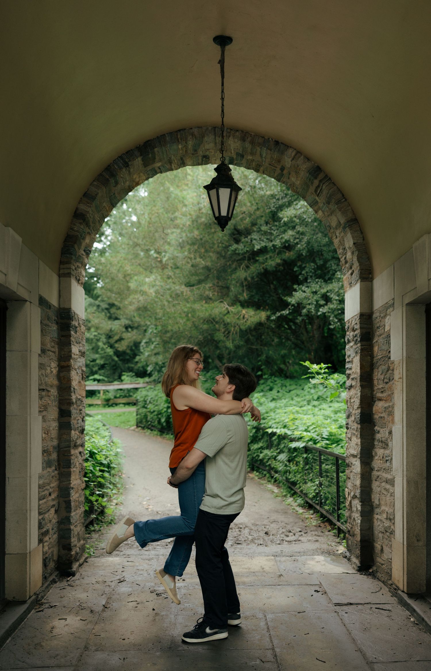 A couple shares a romantic lift embrace framed by a stone archway with lush greenery beyond.