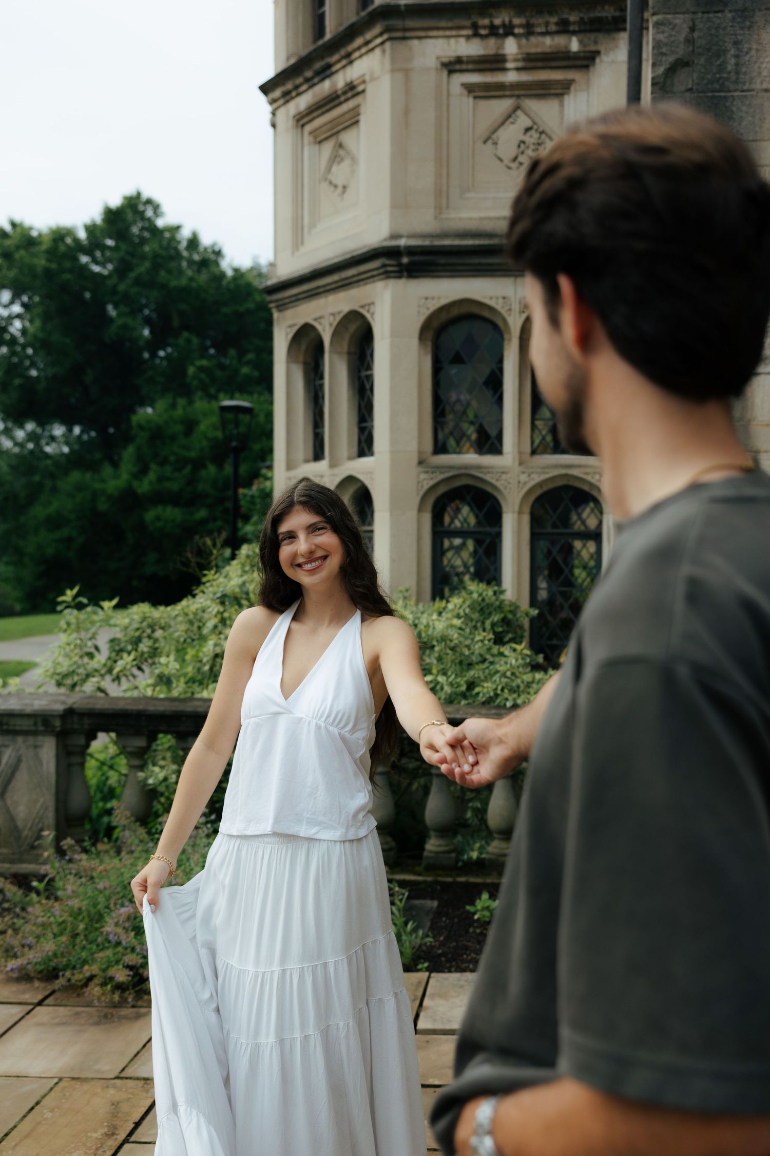 Smiling brunette woman in white maxi dress holds mans hand near historic stone building outdoors.