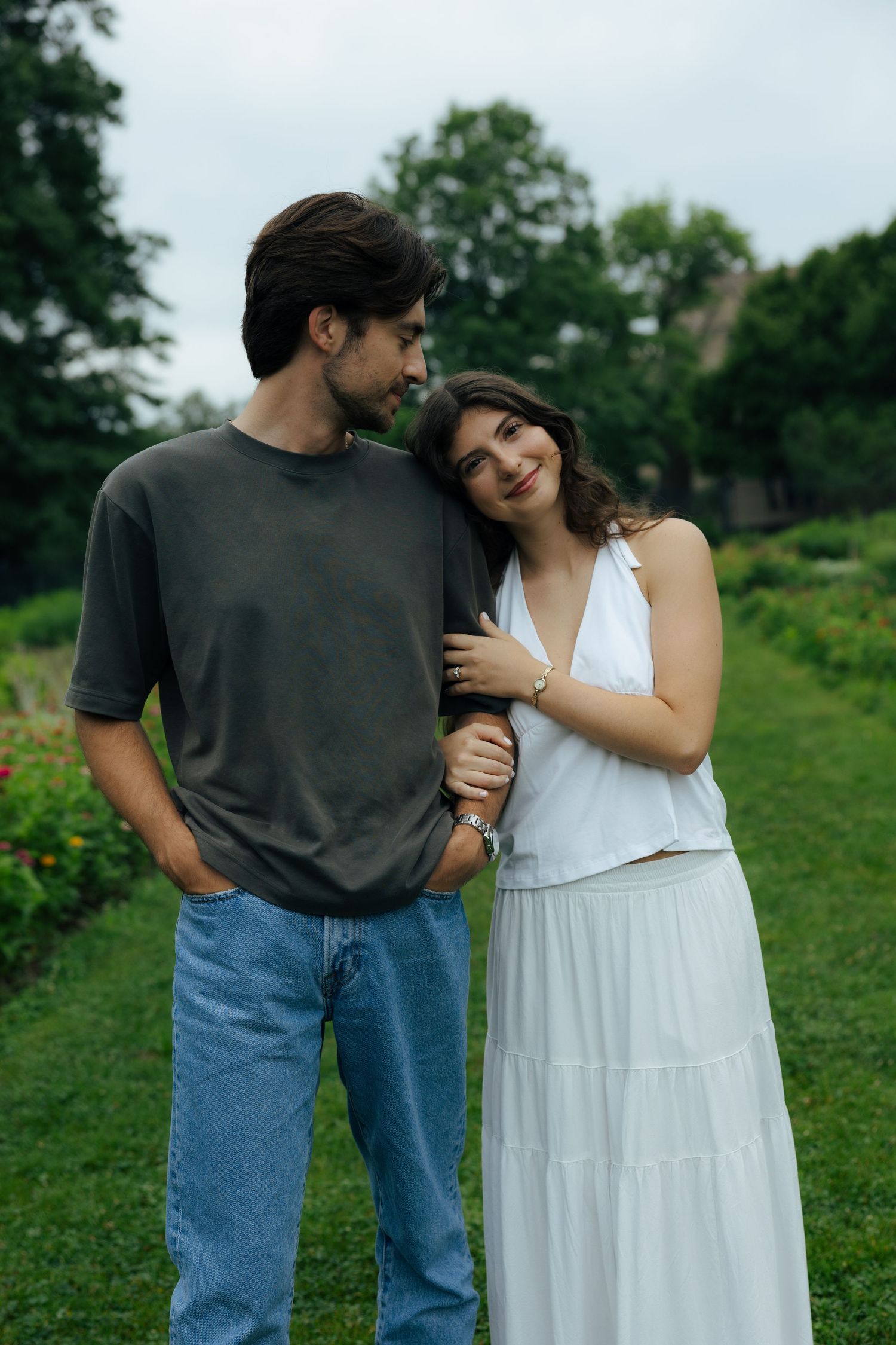 Engaged couple stands close together in a garden, woman in white dress leaning on mans shoulder affectionately.
