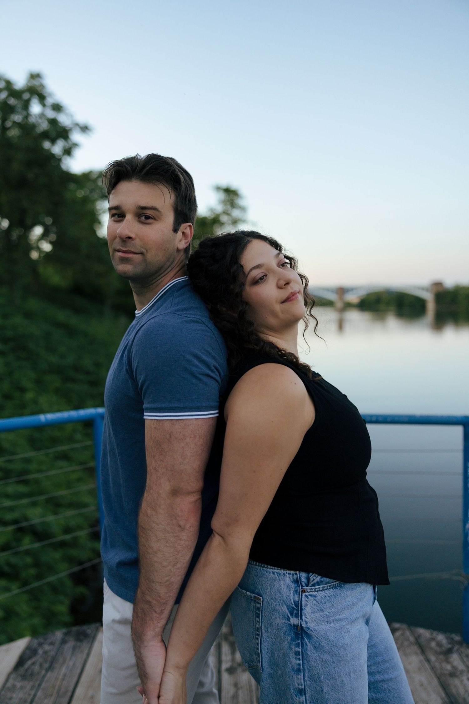 Couple standing back-to-back on a waterfront dock at dusk, holding hands and smiling at the camera.