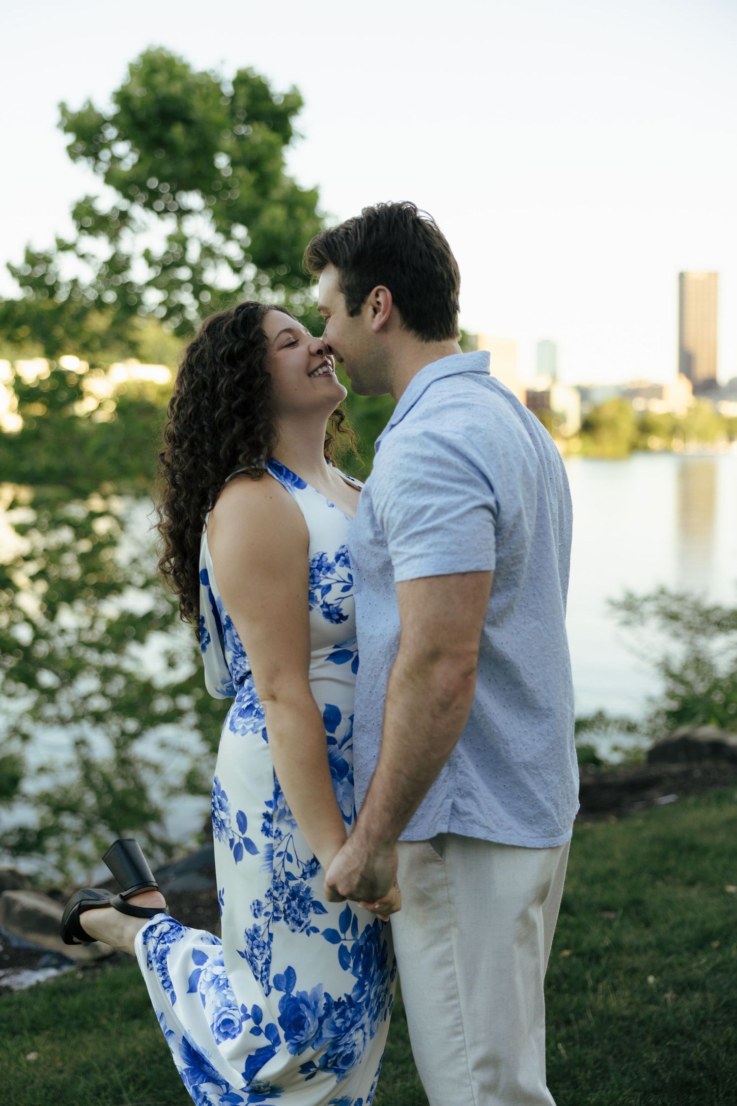 Laughing couple nearly kissing while holding hands near river with city skyline backdrop.
