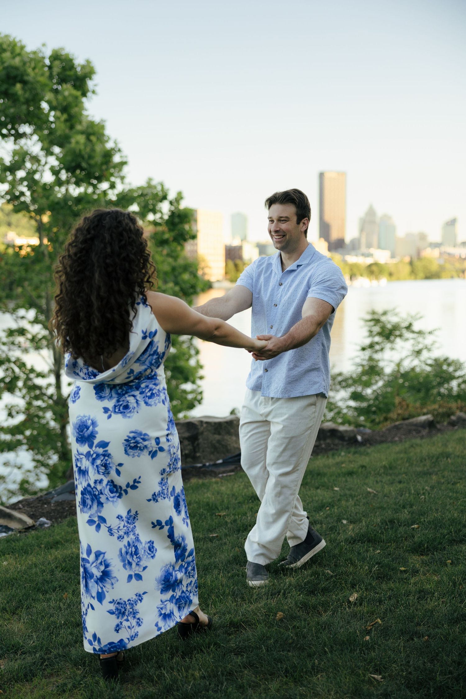 A couple dances and holds hands on a grassy riverbank with a city skyline in the background.