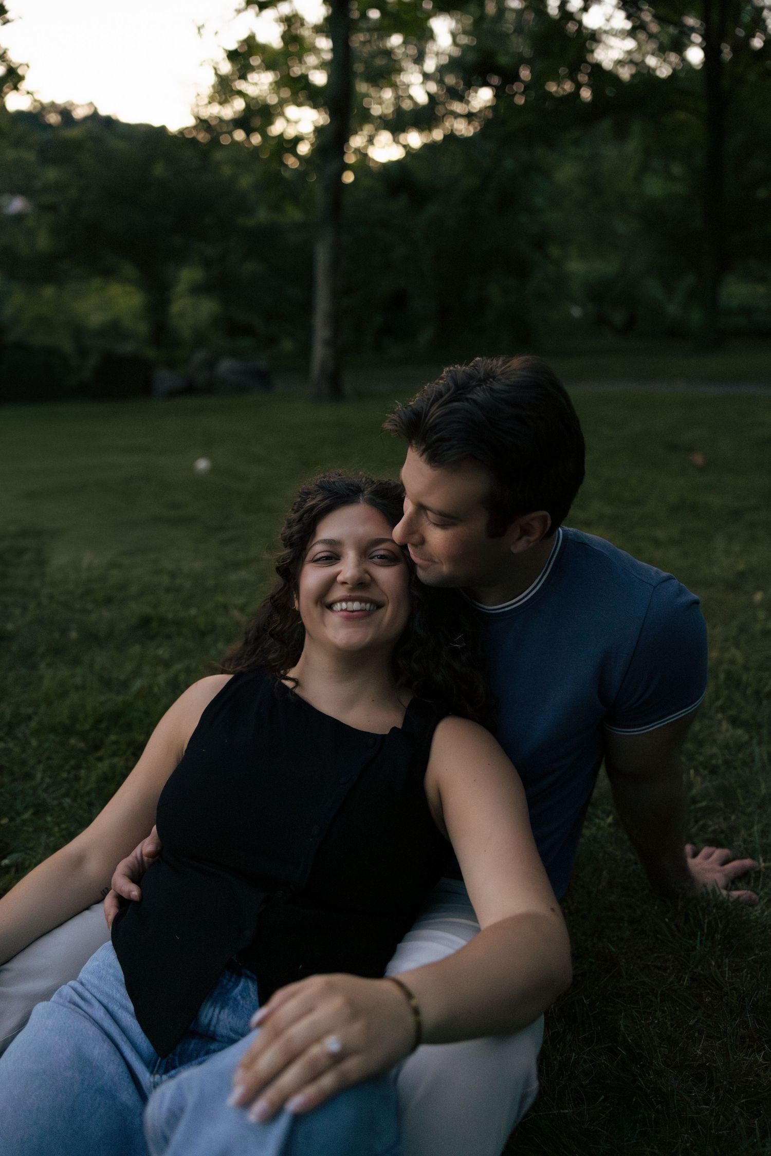 Man kissing woman on cheek as they sit together on grass in park at dusk, woman smiling.