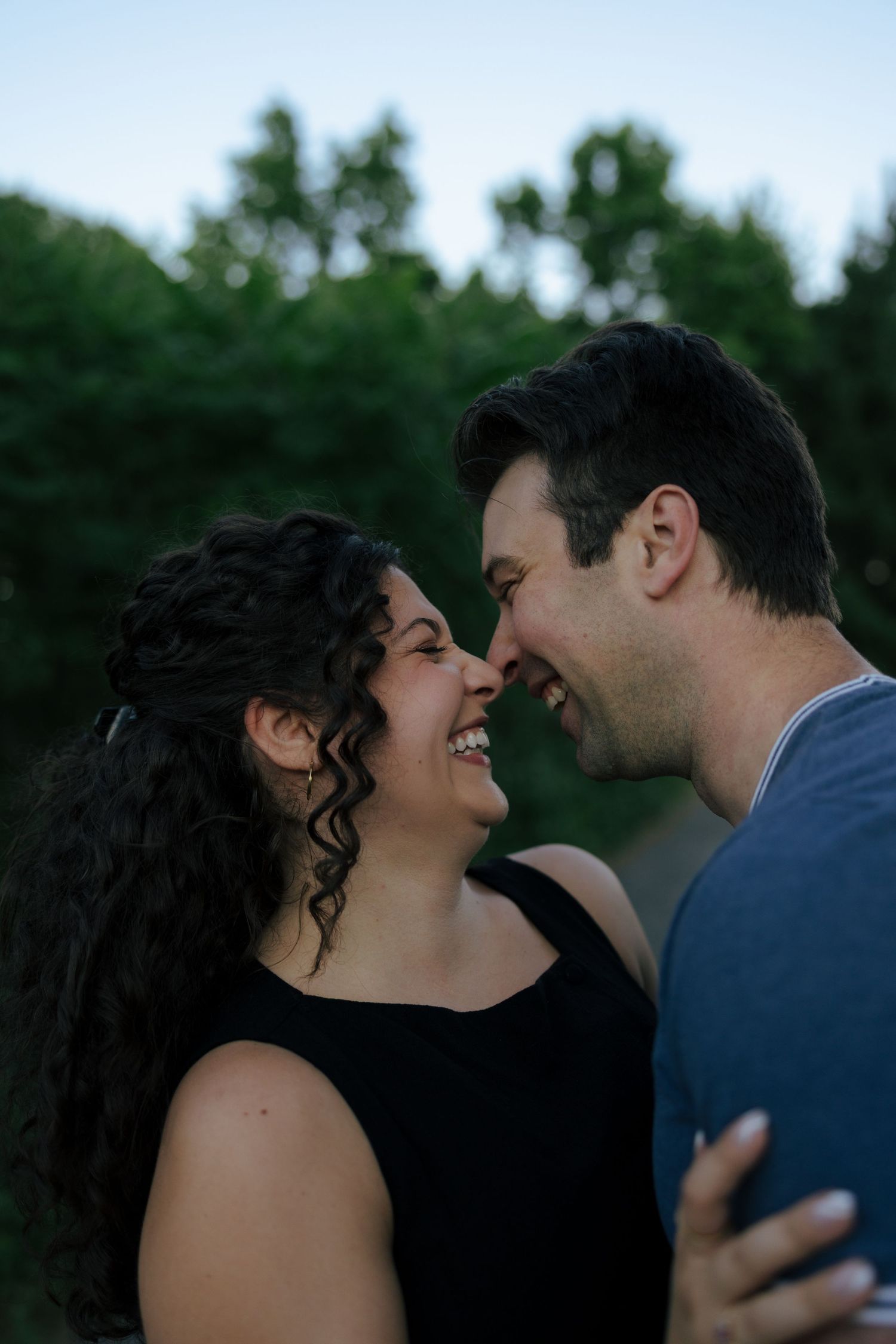 A joyful couple smiles and touches noses outdoors with lush green trees in the background at dusk.
