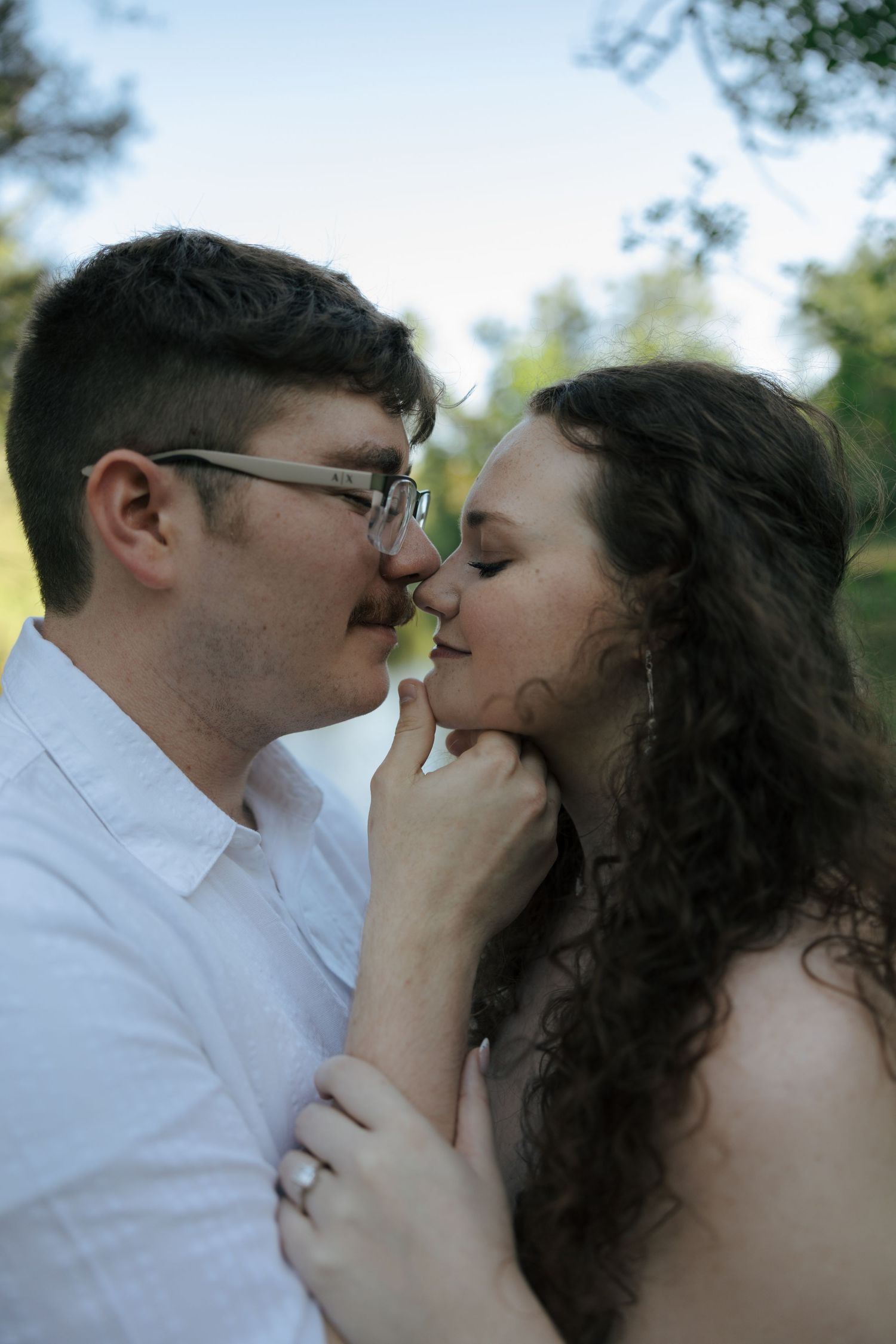 Couple sharing a romantic almost-kiss outdoors, man in white shirt with glasses, woman with curly dark hair, green trees behind.