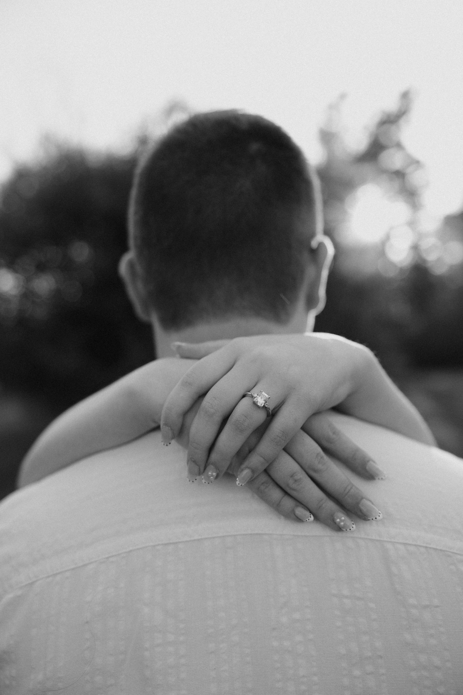 Black and white close-up of a woman's hand with an engagement ring resting on a man's shoulder.
