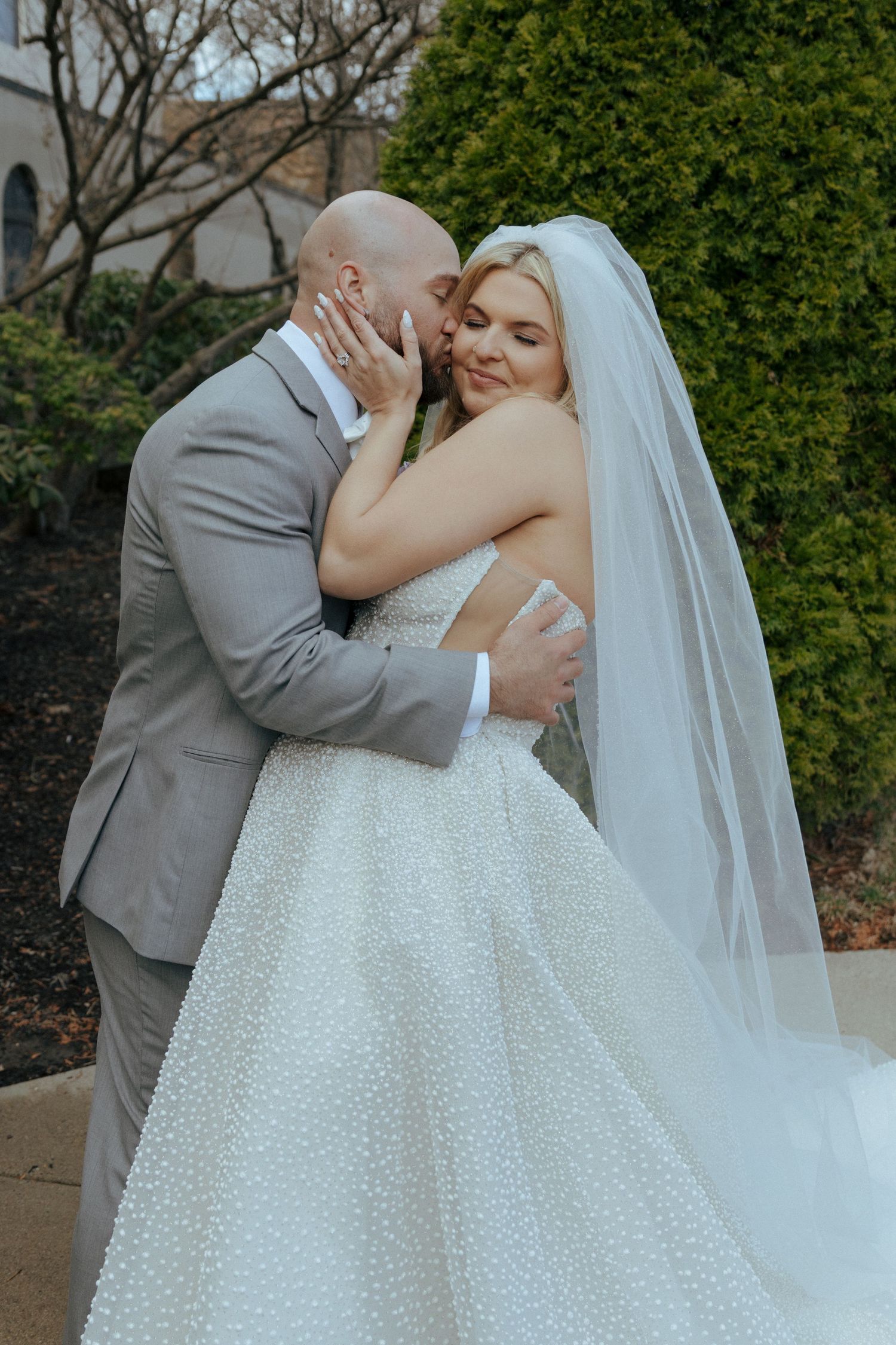 Bride and groom sharing a romantic kiss outdoors, bride in a white ballgown with veil, groom in a gray suit.