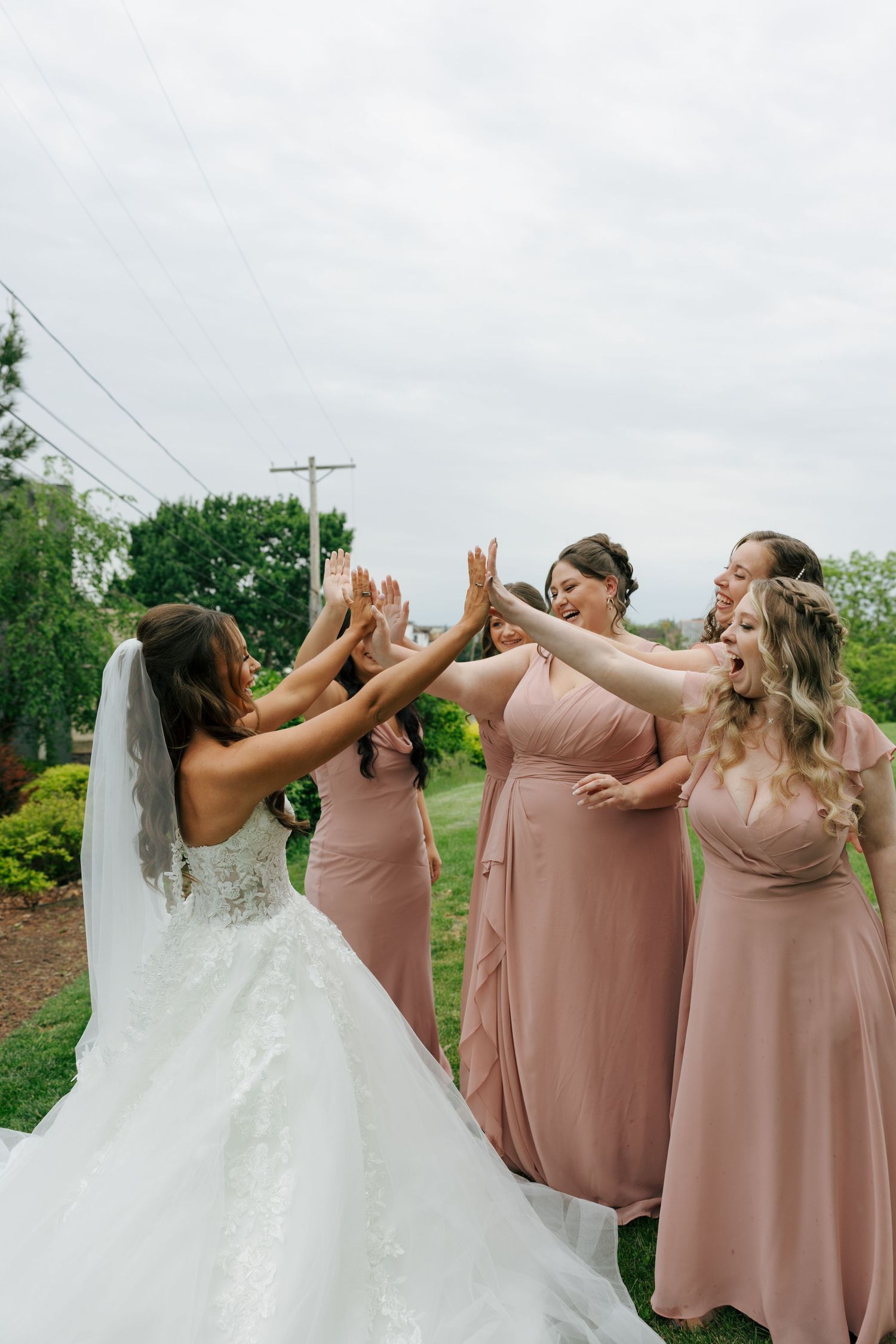 Bride and bridesmaids in blush pink gowns joyfully high-fiving outdoors on a cloudy wedding day.