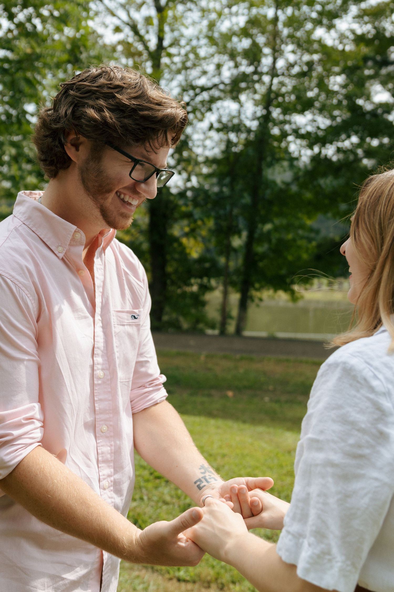 A couple in pink and white shirts playfully dance together in a sunny outdoor park setting.