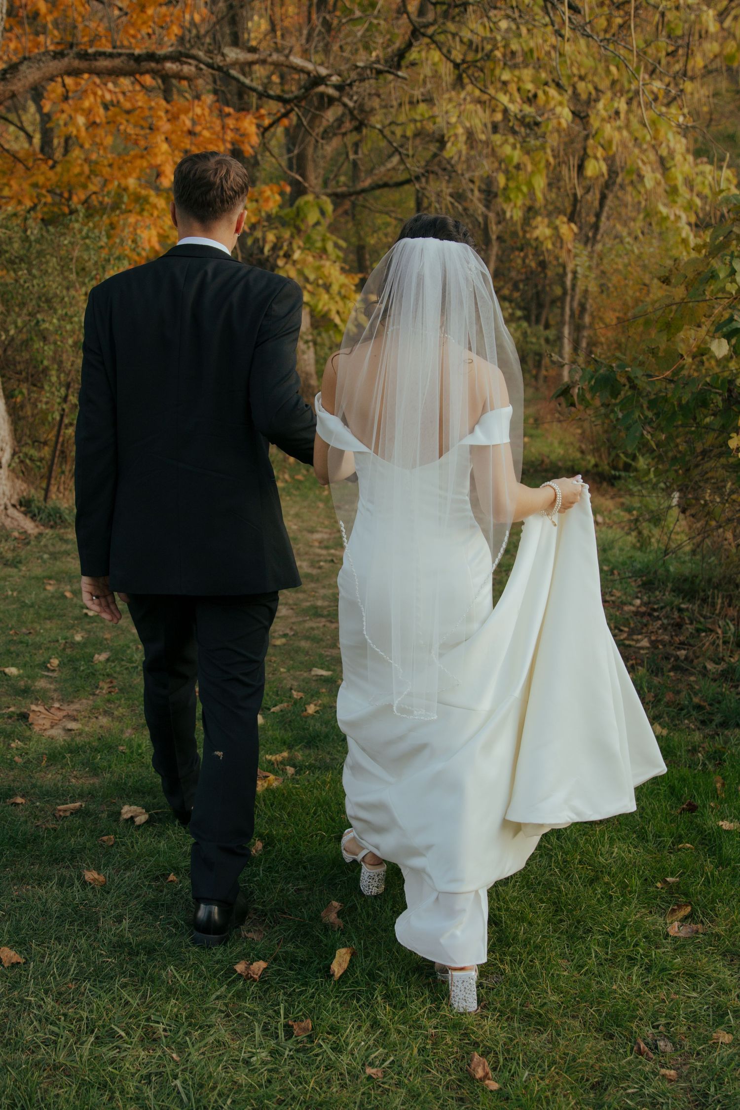 A couple in wedding attire walks through autumn foliage as the bride's white veil flows behind them in the breeze.