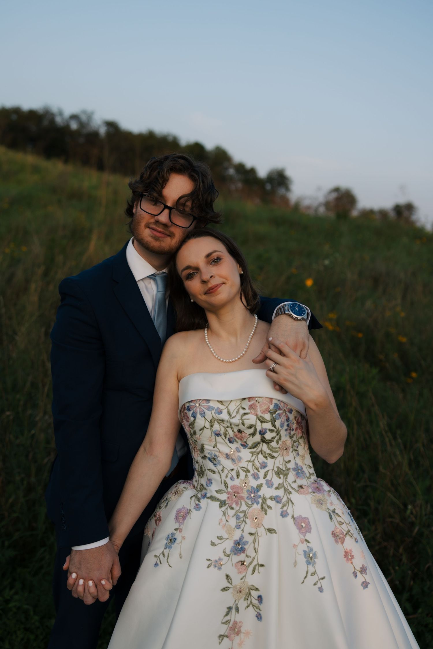 Bride in floral embroidered strapless gown and groom in navy suit embrace in a green meadow at dusk.