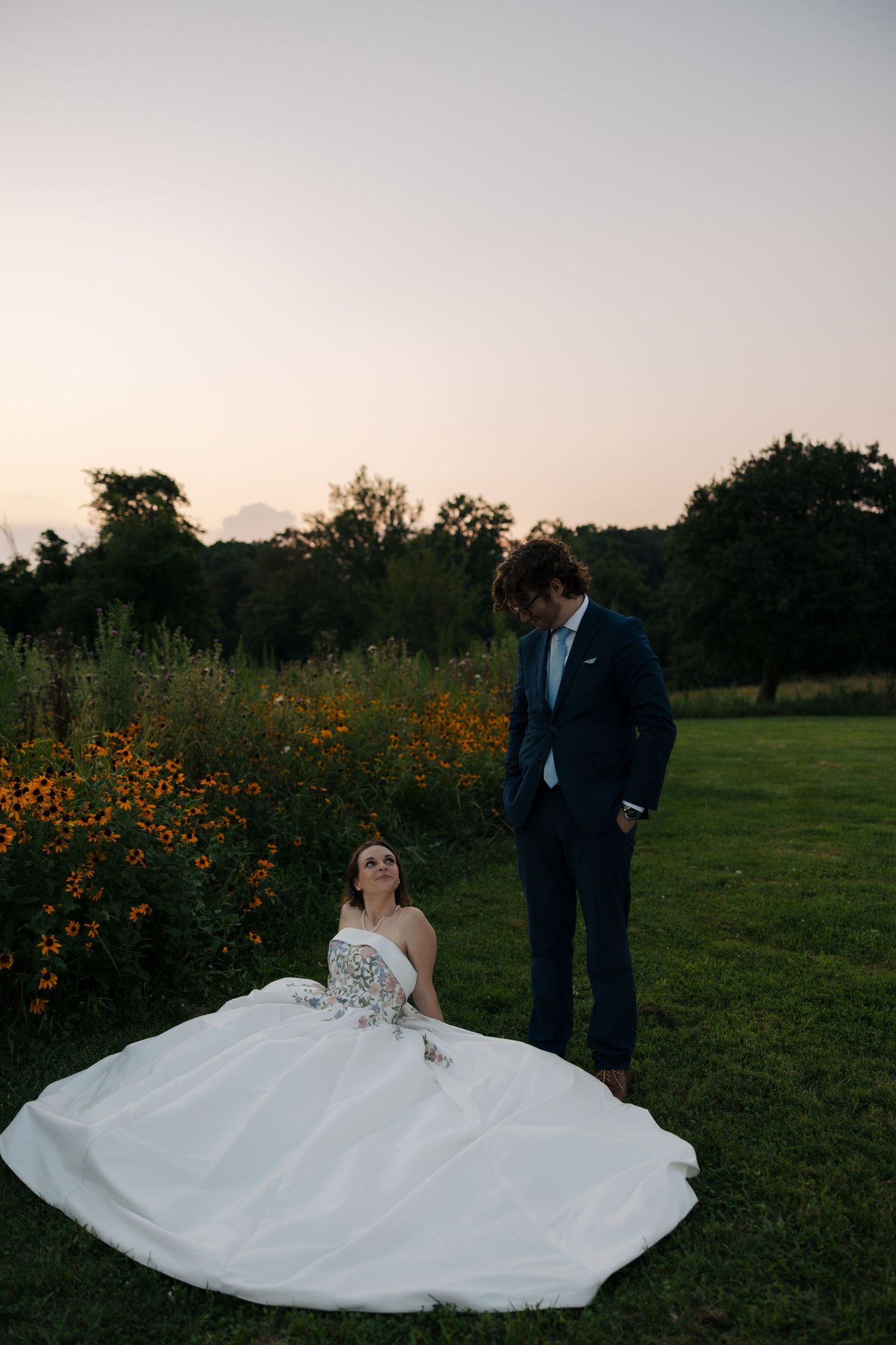 Bride in strapless floral gown sits by wildflowers as groom in navy suit stands nearby.