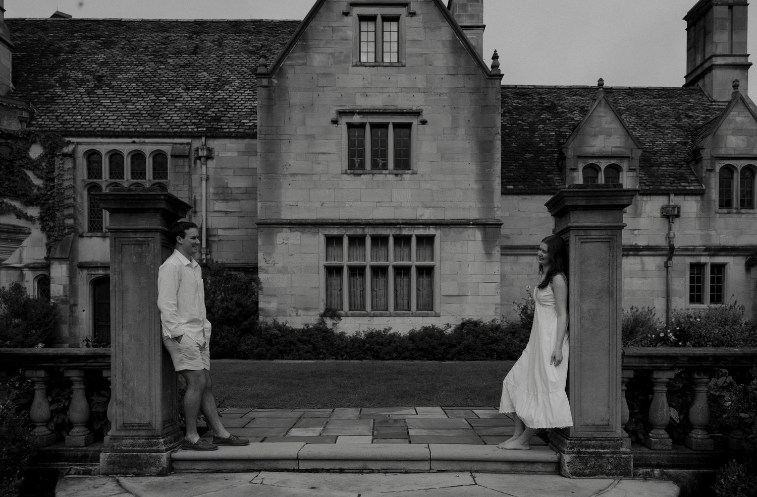 Black and white photo of couple standing between stone columns before a historic English estate.