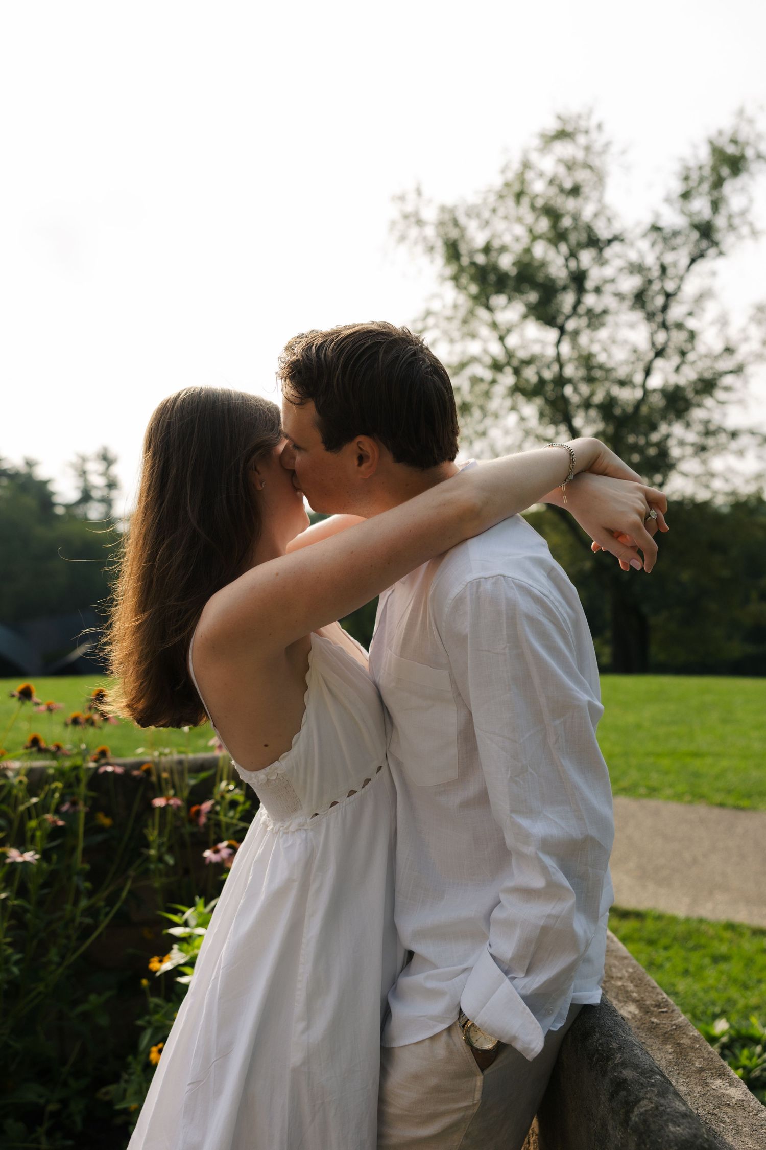 Couple in white outfits sharing a romantic kiss outdoors in a lush green garden setting.