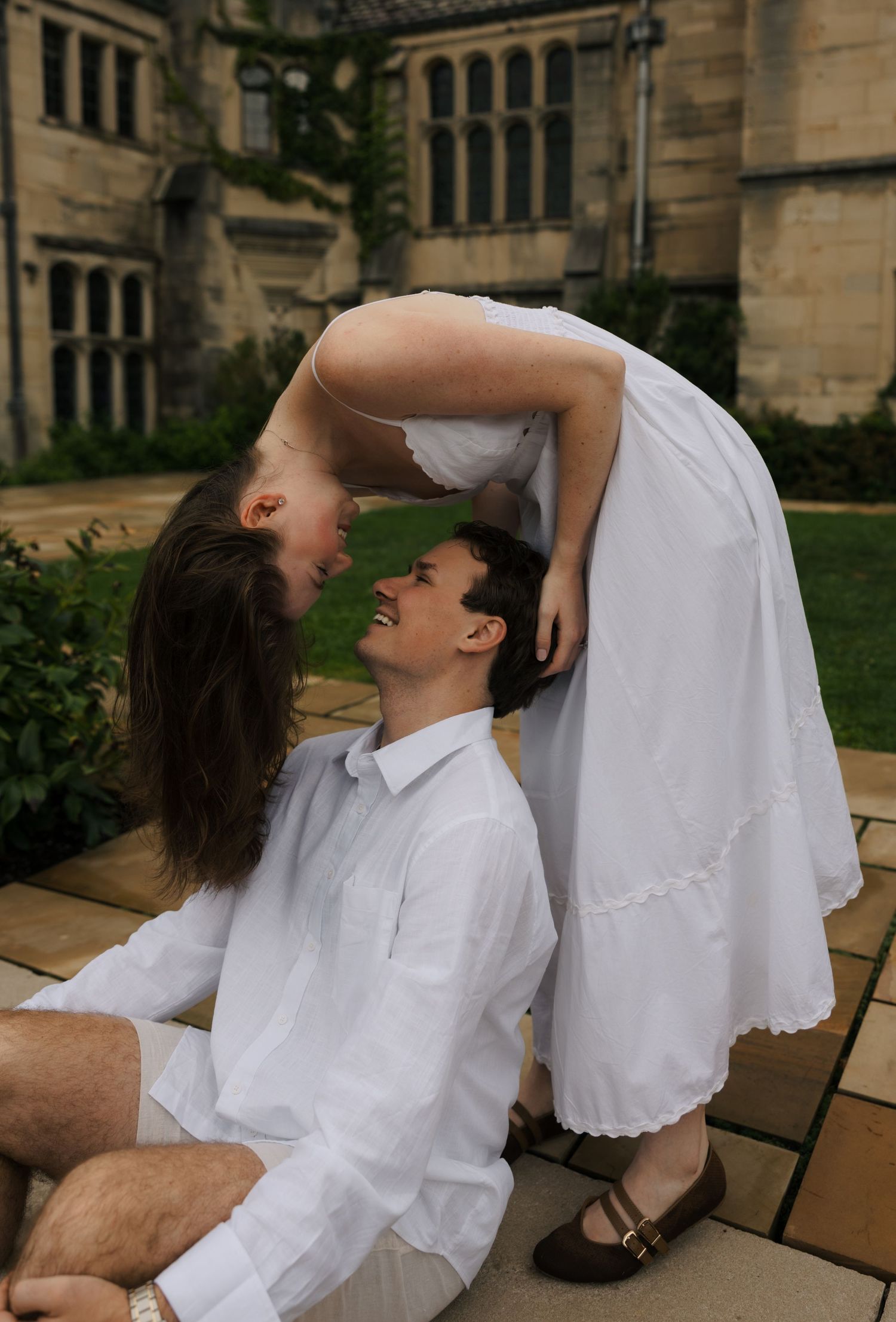 Joyful couple in white clothing sharing a playful moment, woman leaning over laughing man seated on stone steps.