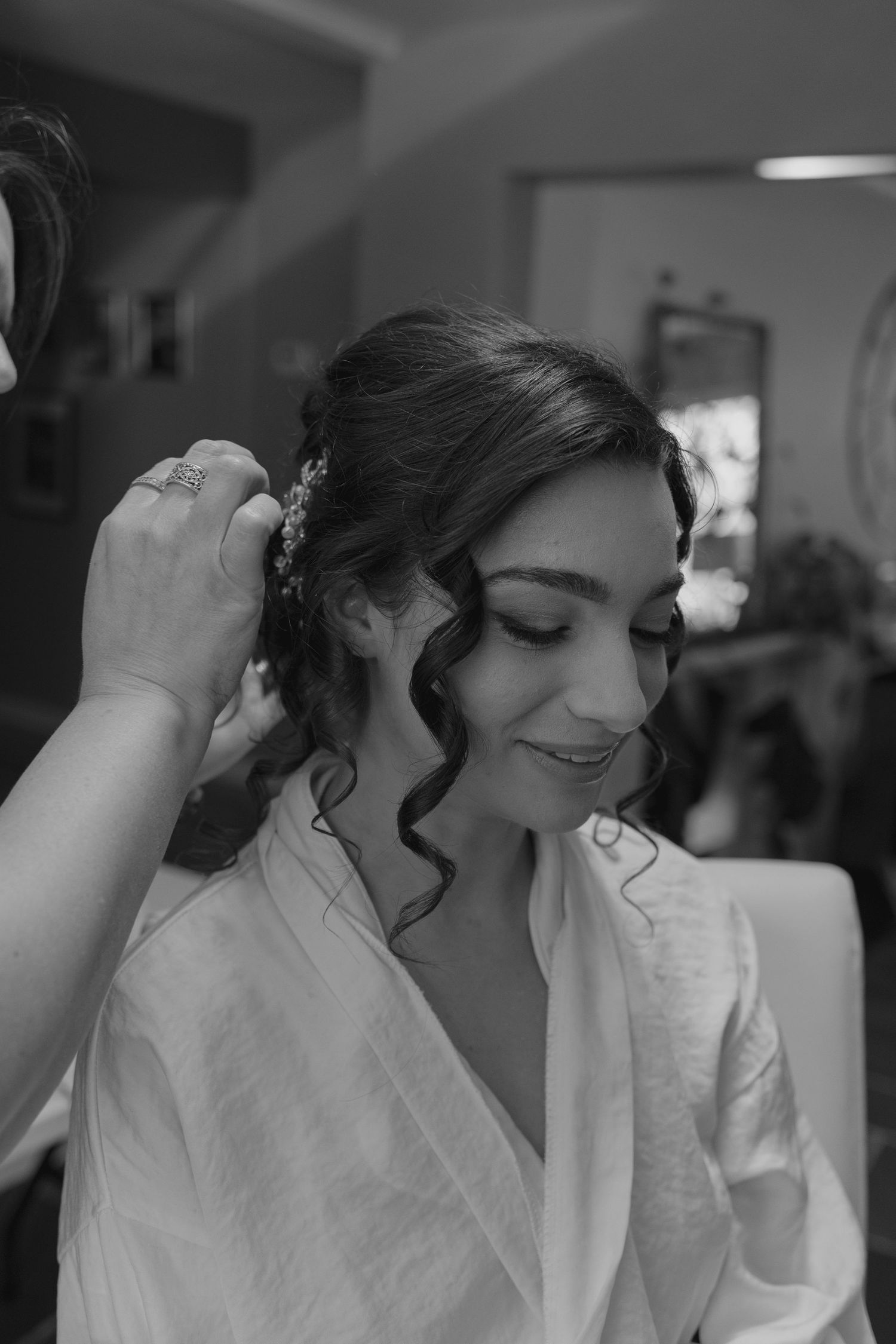 Bride smiling as hairstylist places floral hair accessory in elegant updo with curls on wedding day.