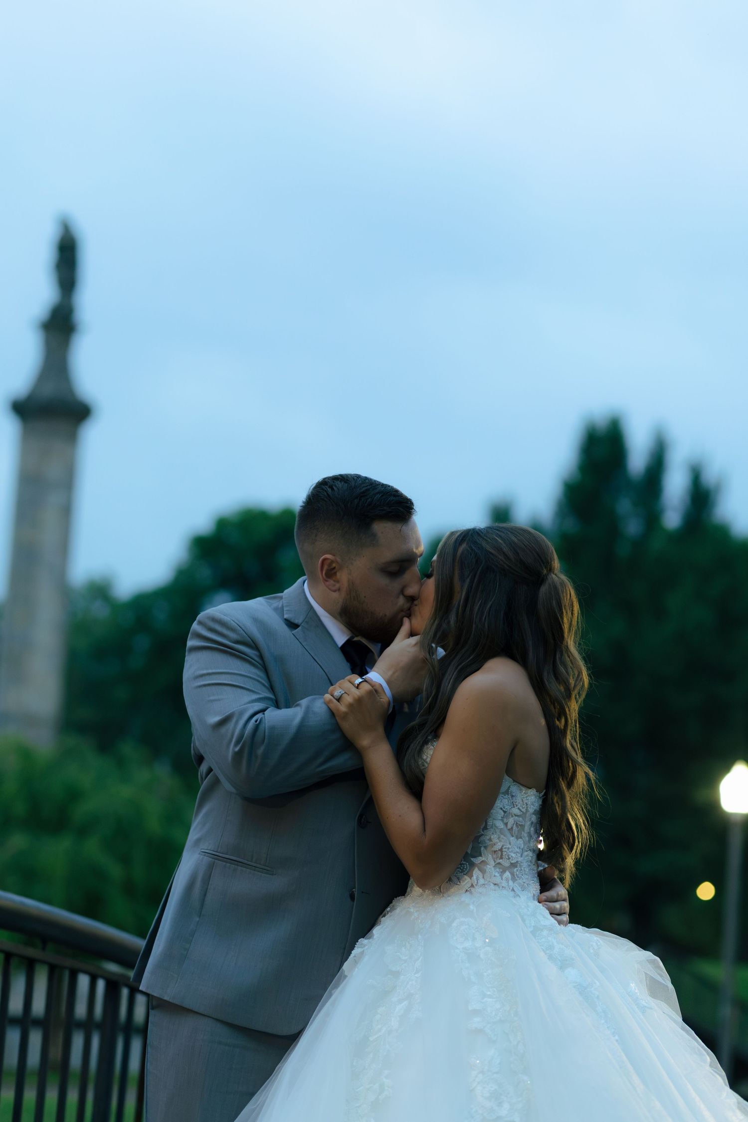 A bride and groom share a romantic kiss outdoors at dusk, with a monument and trees visible in the background.