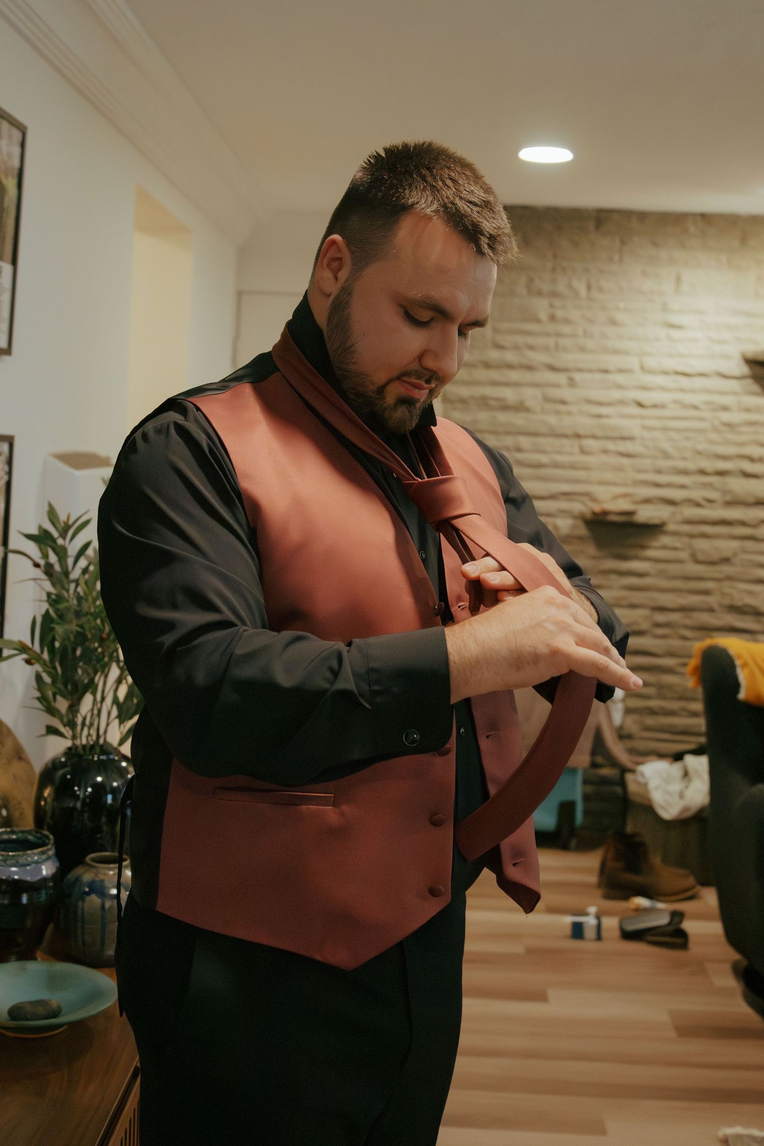 Groom in burgundy vest and black shirt adjusts tie while getting dressed for wedding ceremony.