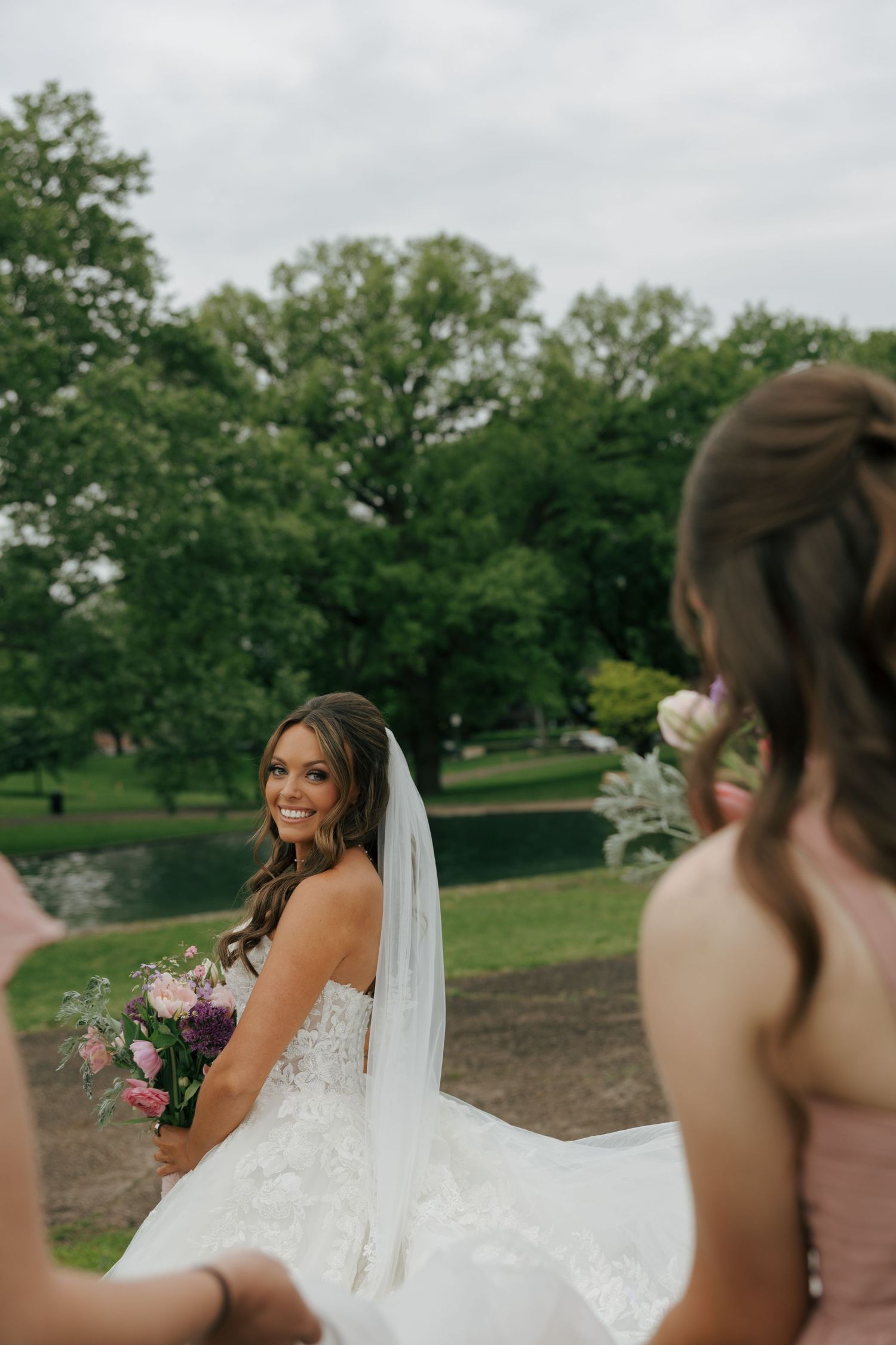 Smiling bride in white lace gown holds pink bouquet while bridesmaids look on outdoors.