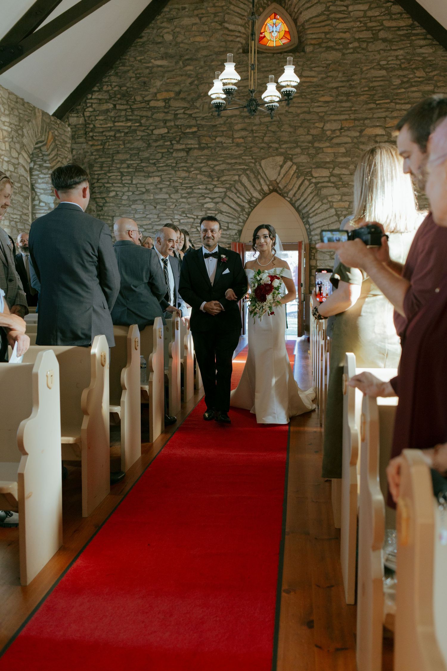 Wedding processional in rustic stone church with red carpet aisle and wooden pews.