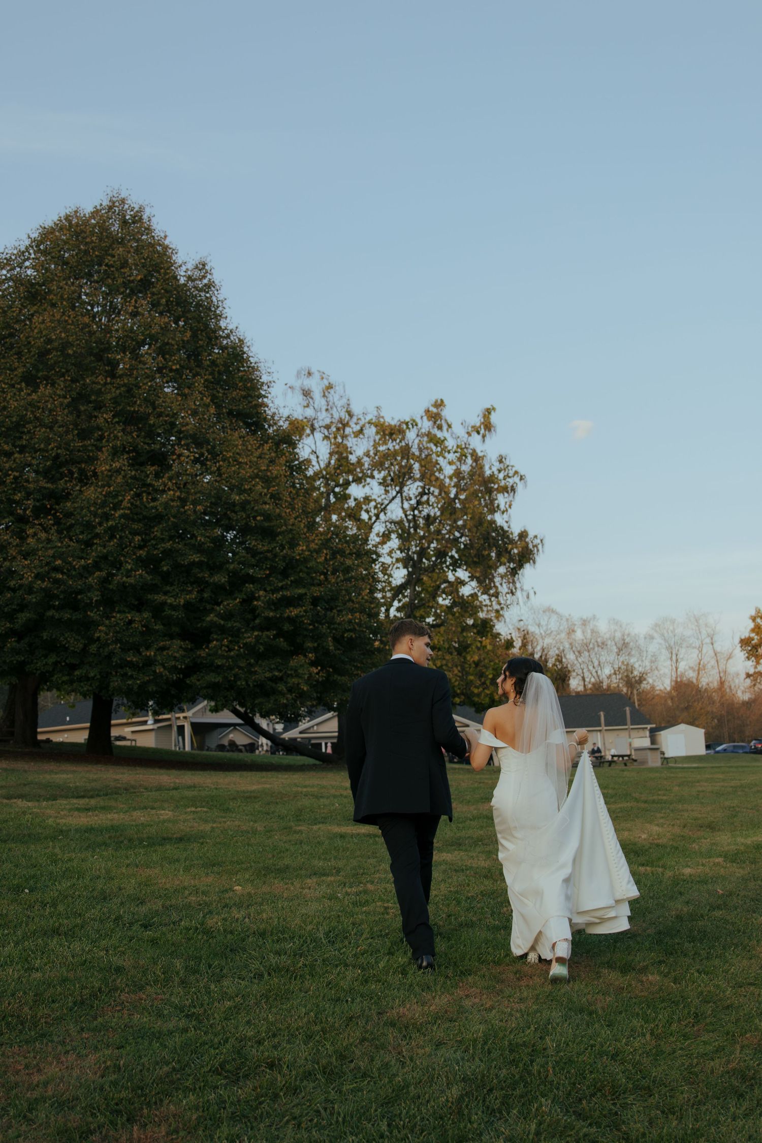 Bride and groom walking across a green lawn at dusk with trees in background.