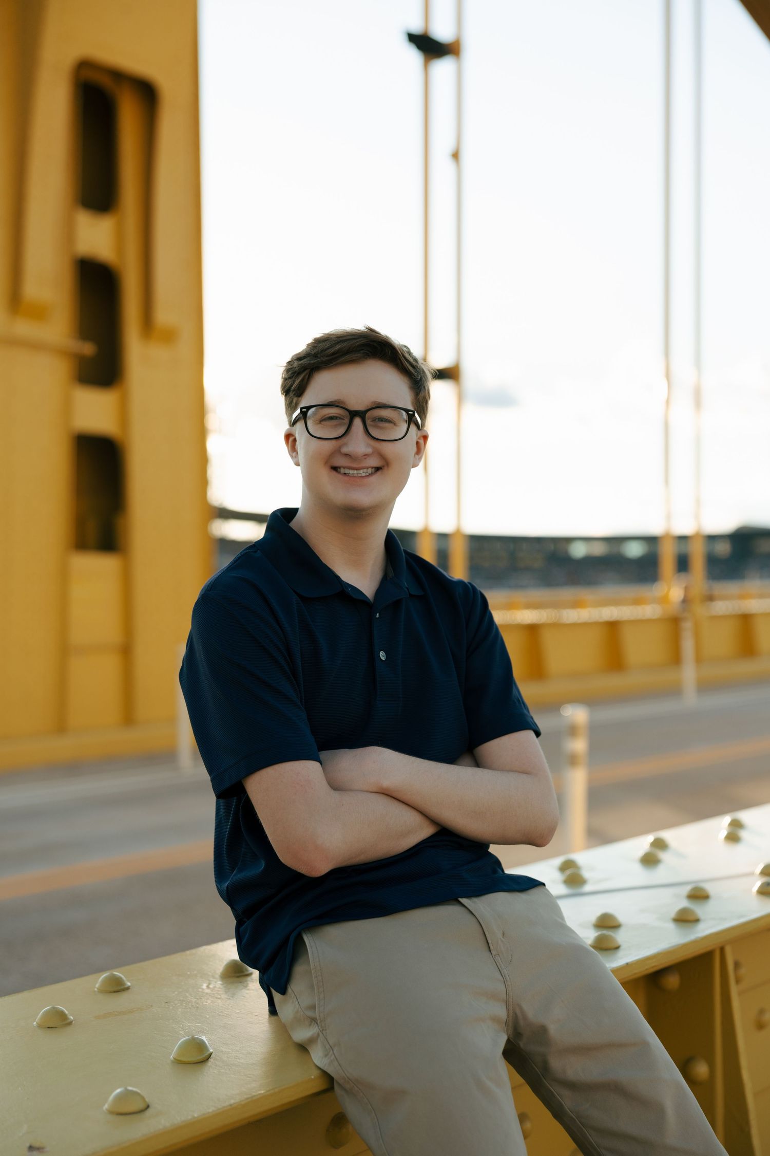 Smiling teen boy in navy polo crosses arms while leaning on yellow bridge railing outdoors.