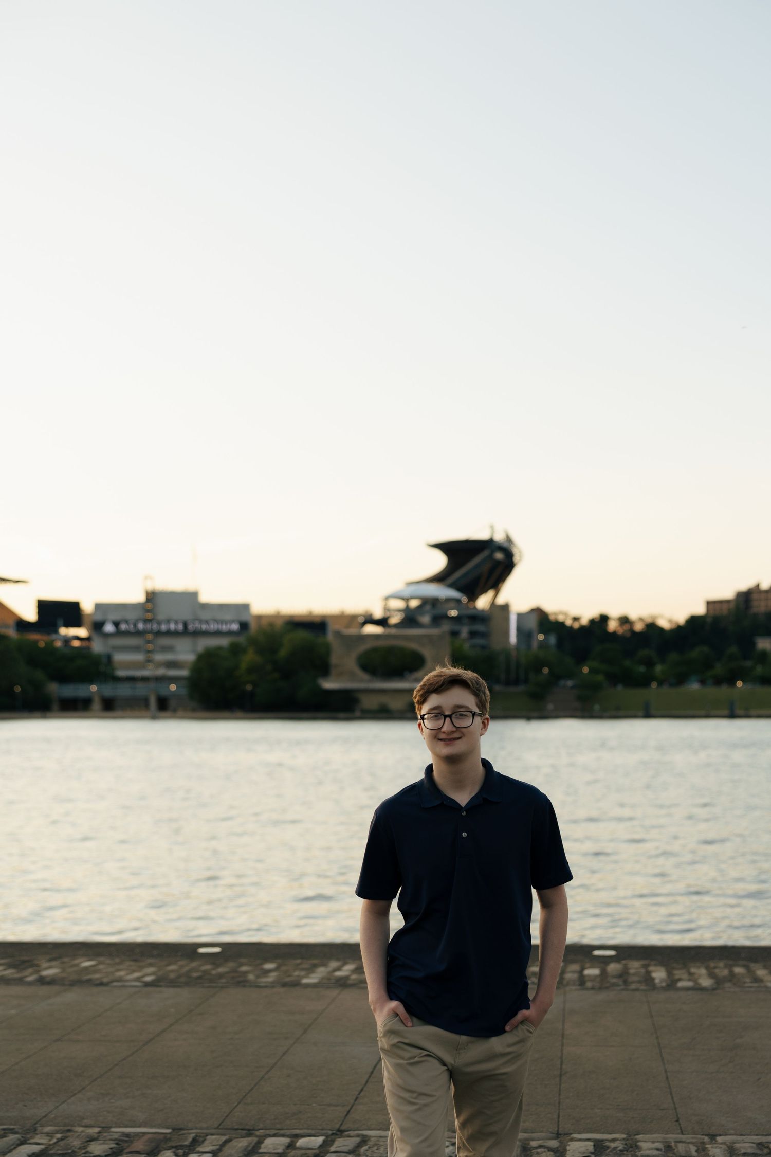 Teen boy in navy polo and glasses stands by a river with a stadium visible in the background.