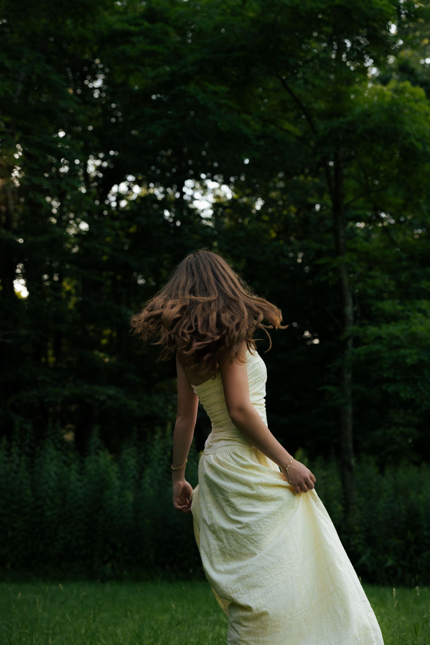 Woman in flowing yellow dress spinning in lush green forest clearing, hair swirling, viewed from behind.