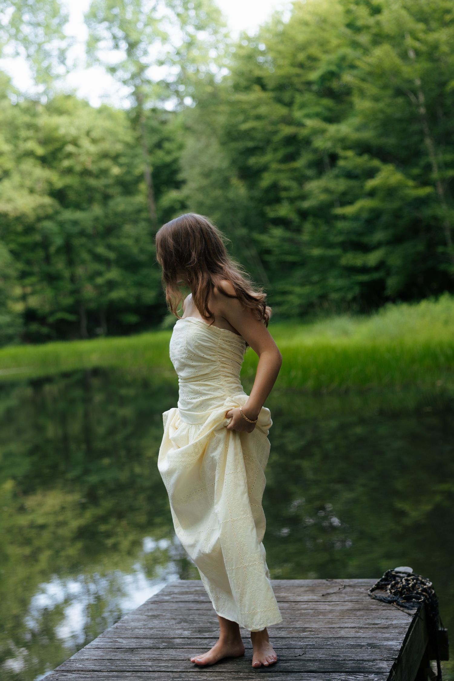 Barefoot woman in cream strapless gown standing on wooden dock overlooking calm forest pond.