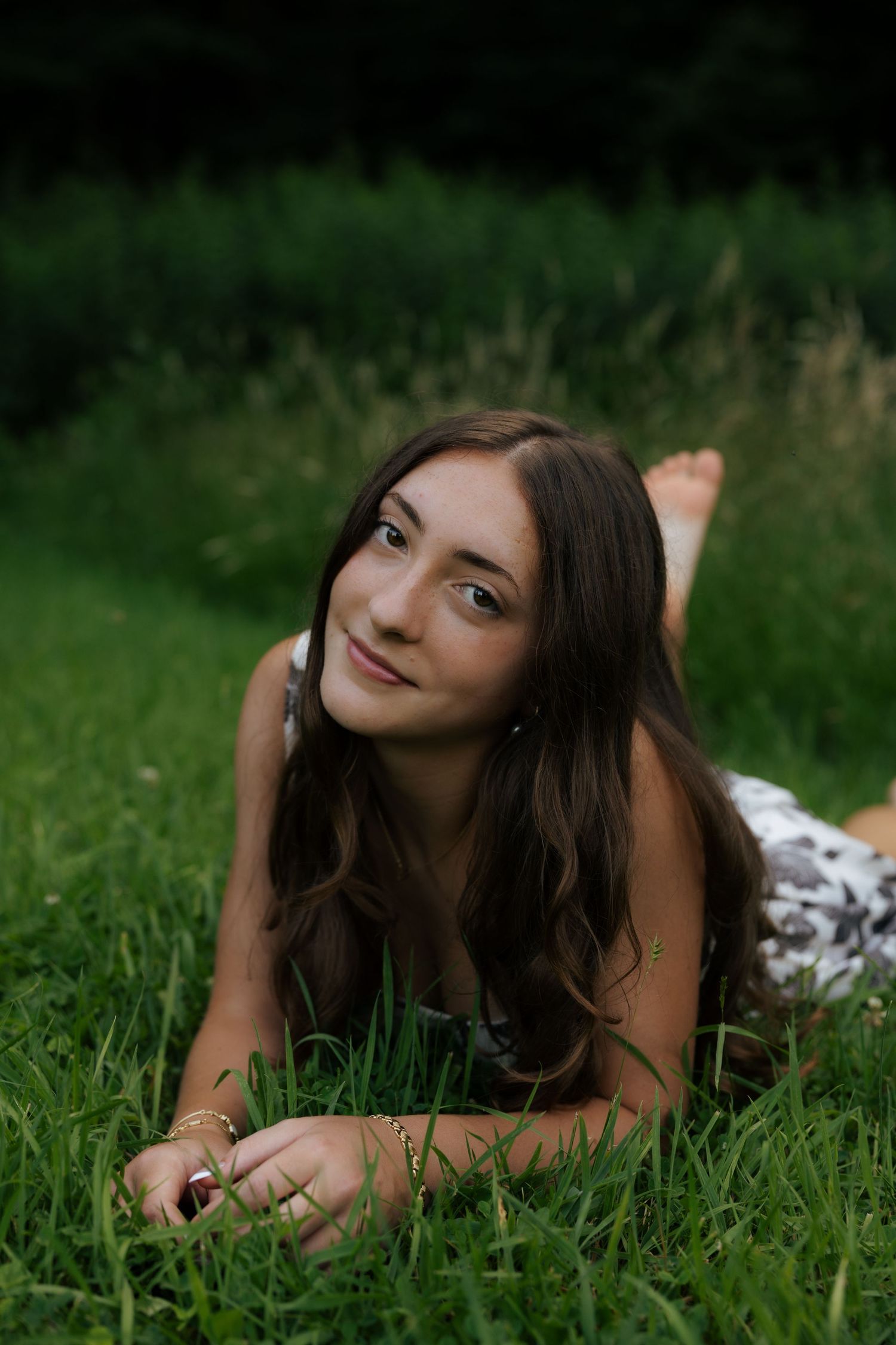 Smiling young woman with long brown hair lying on green grass wearing a floral dress outdoors.