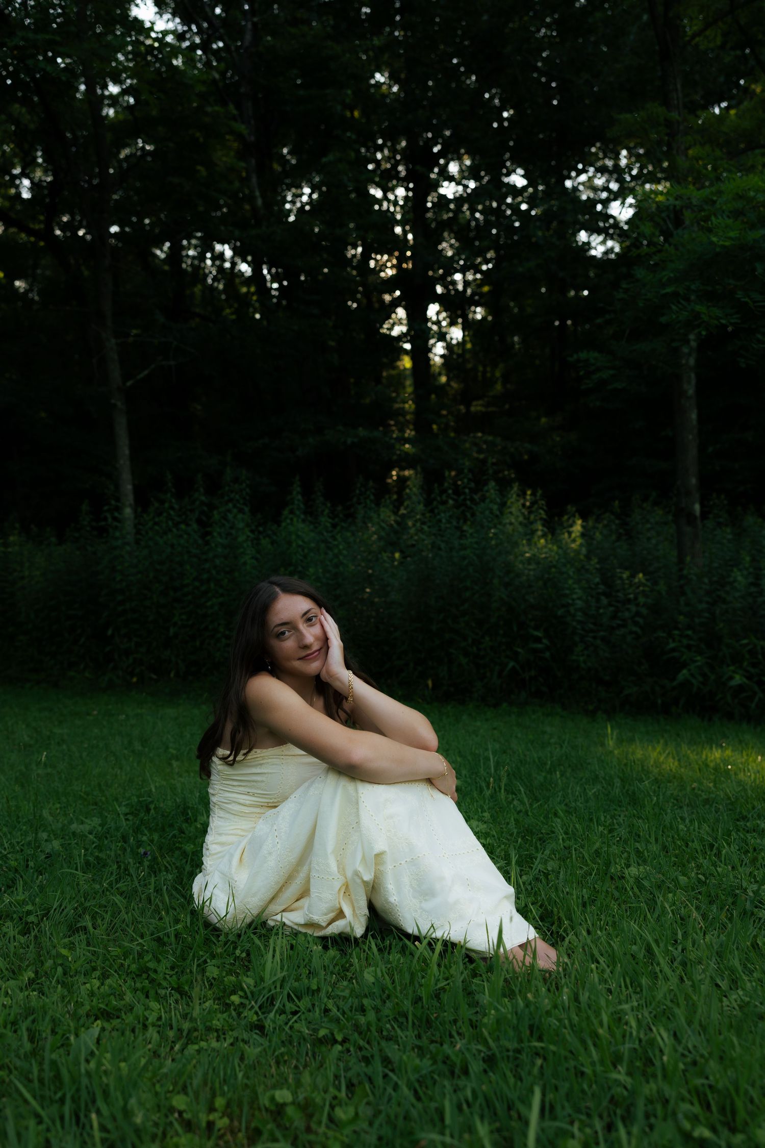 Young woman in white dress sitting on grass smiling with dark forest background at dusk.