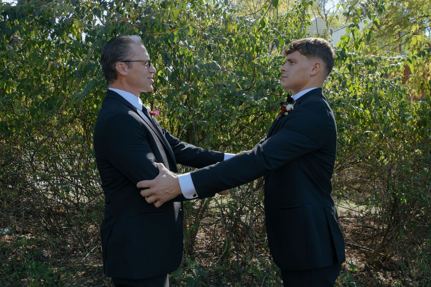 Two men in black tuxedos adjusting each other's suits outdoors with lush green foliage in background.