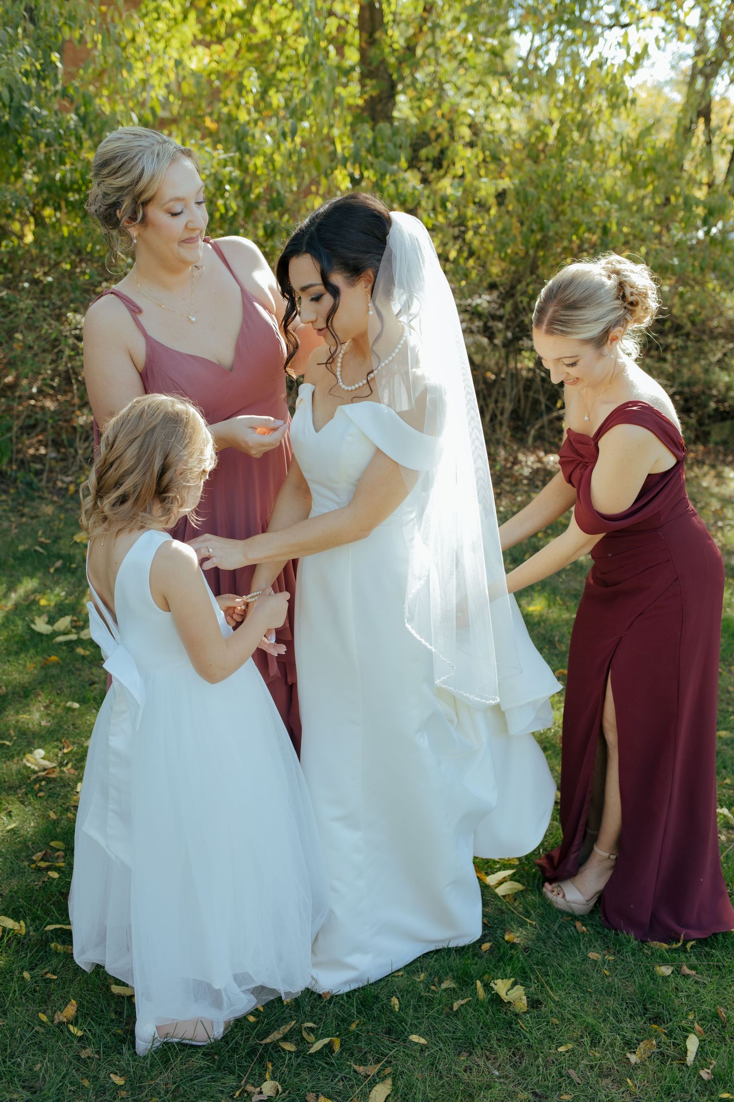 Bride in white gown with veil surrounded by bridesmaids in mauve and burgundy dresses outdoors.