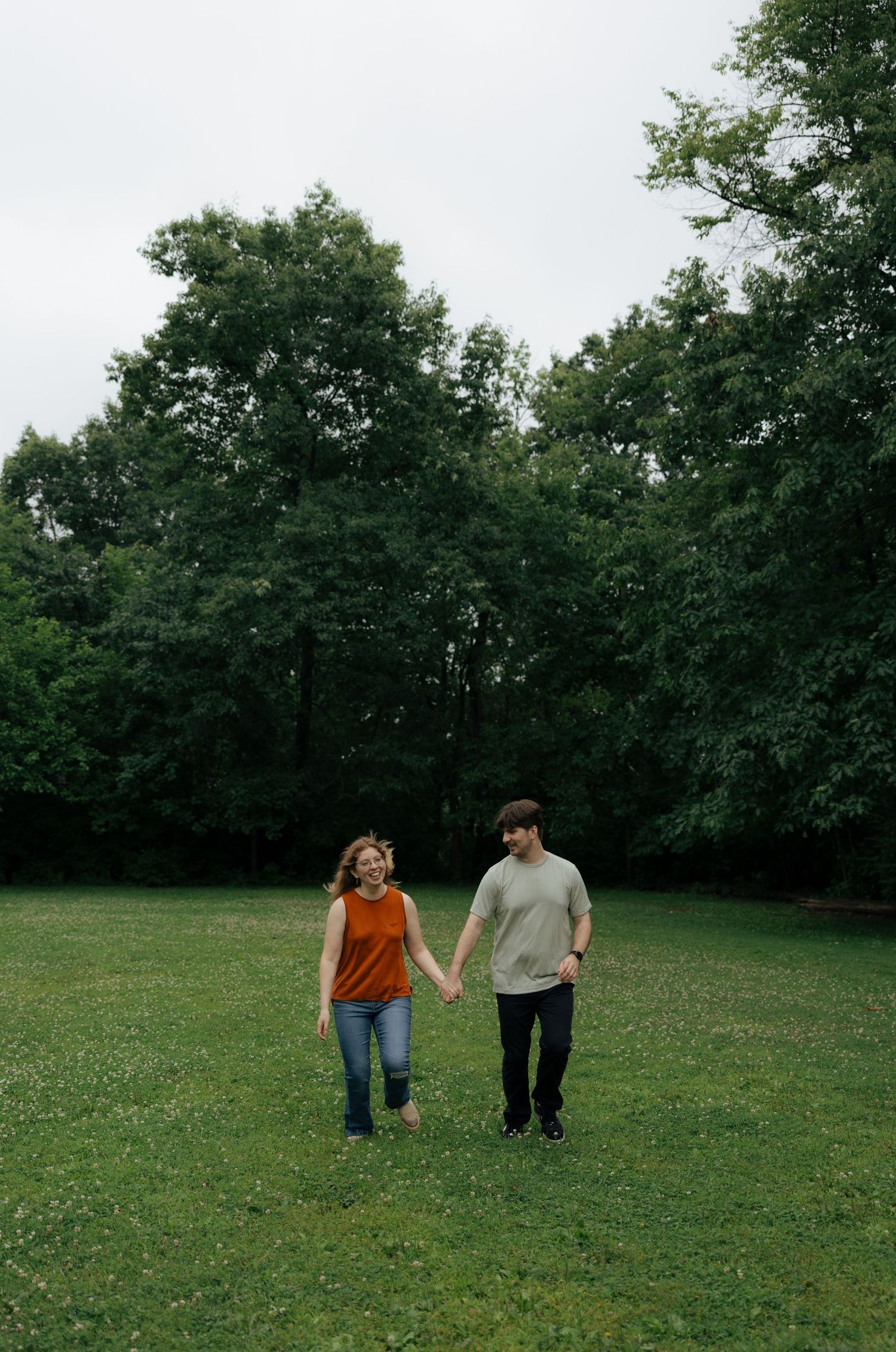 A couple holds hands while walking through a lush green park surrounded by tall trees on an overcast day.