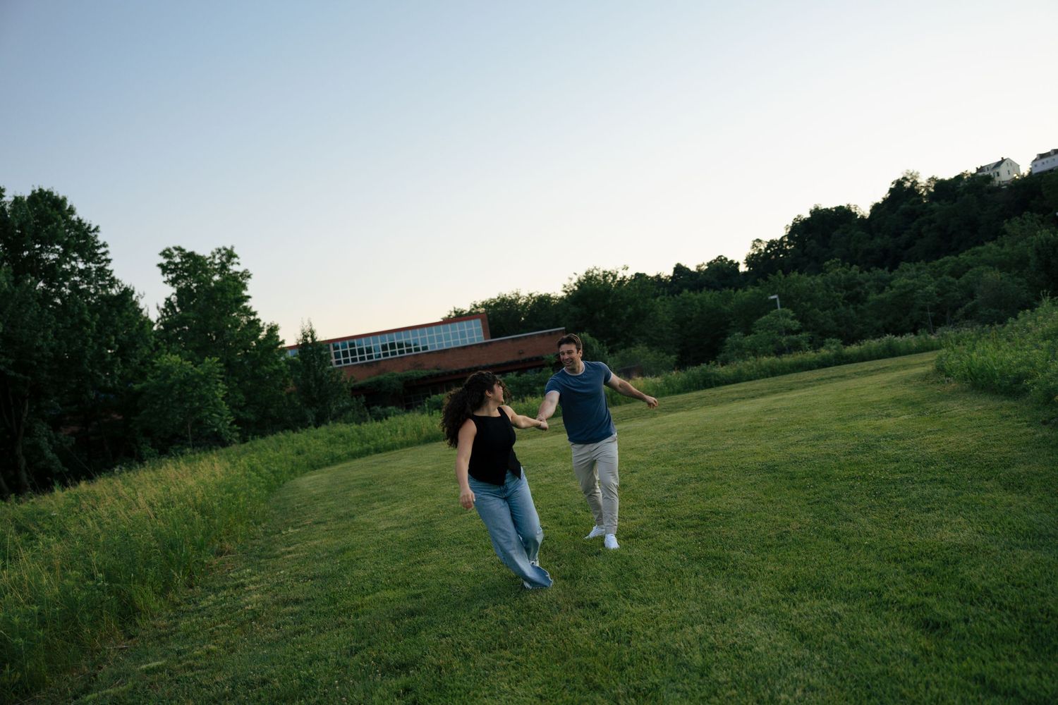 A couple holds hands and runs playfully across a green field with a bridge visible in the background.
