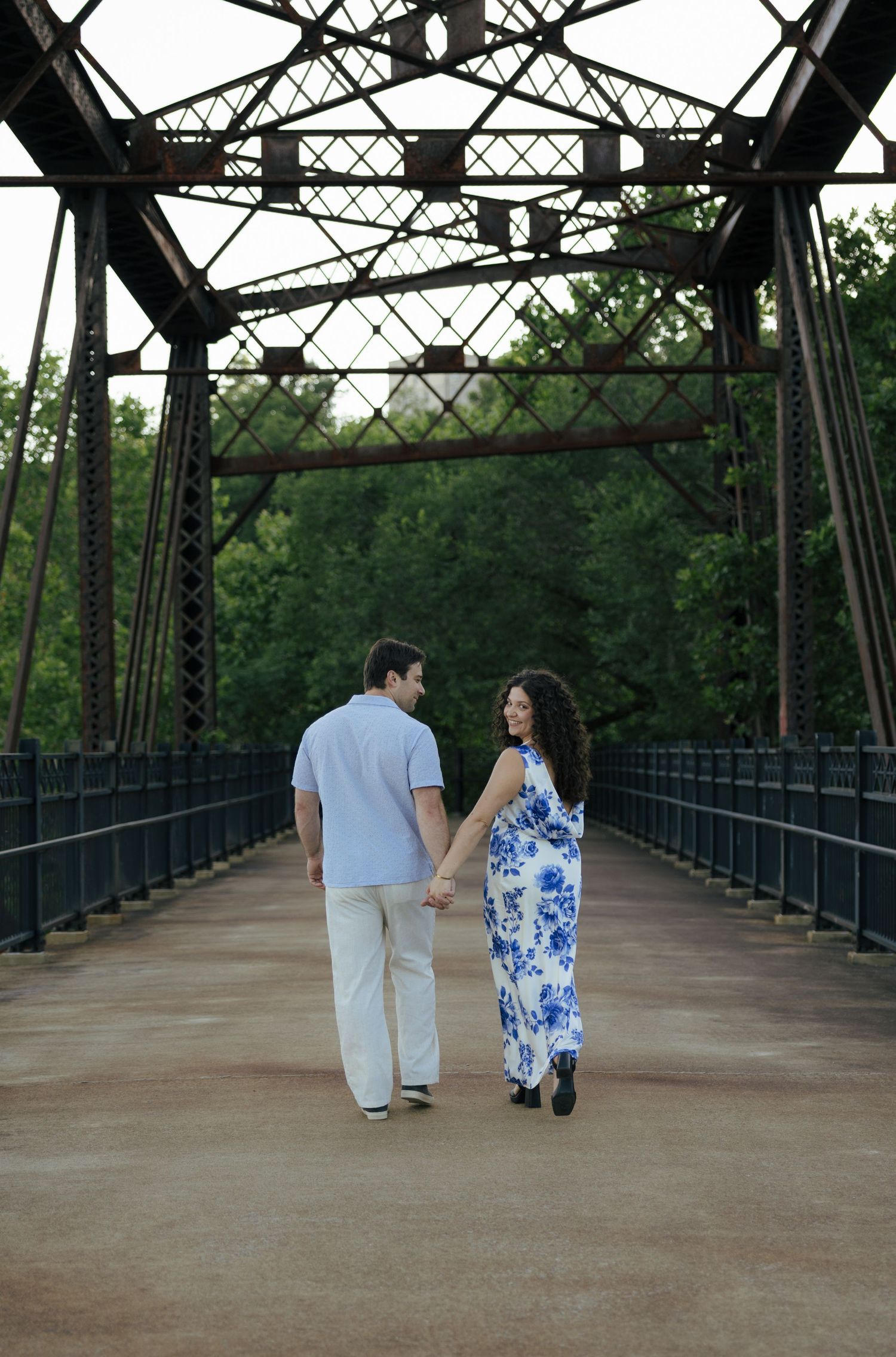 A couple holds hands while strolling away on a vintage iron bridge with green trees visible in the background.