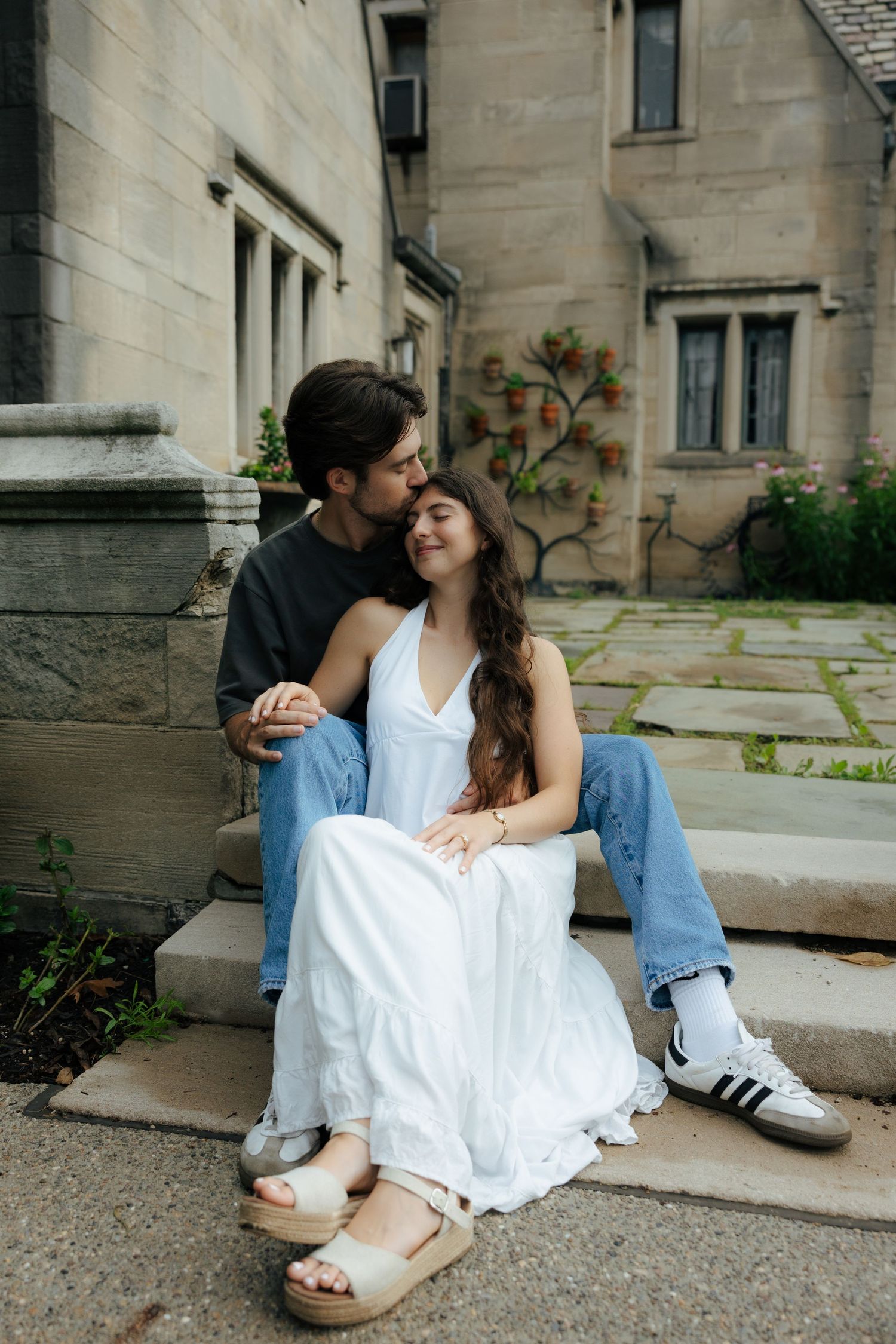 A couple sits together on stone steps outside a historic building, sharing a gentle kiss on the cheek.