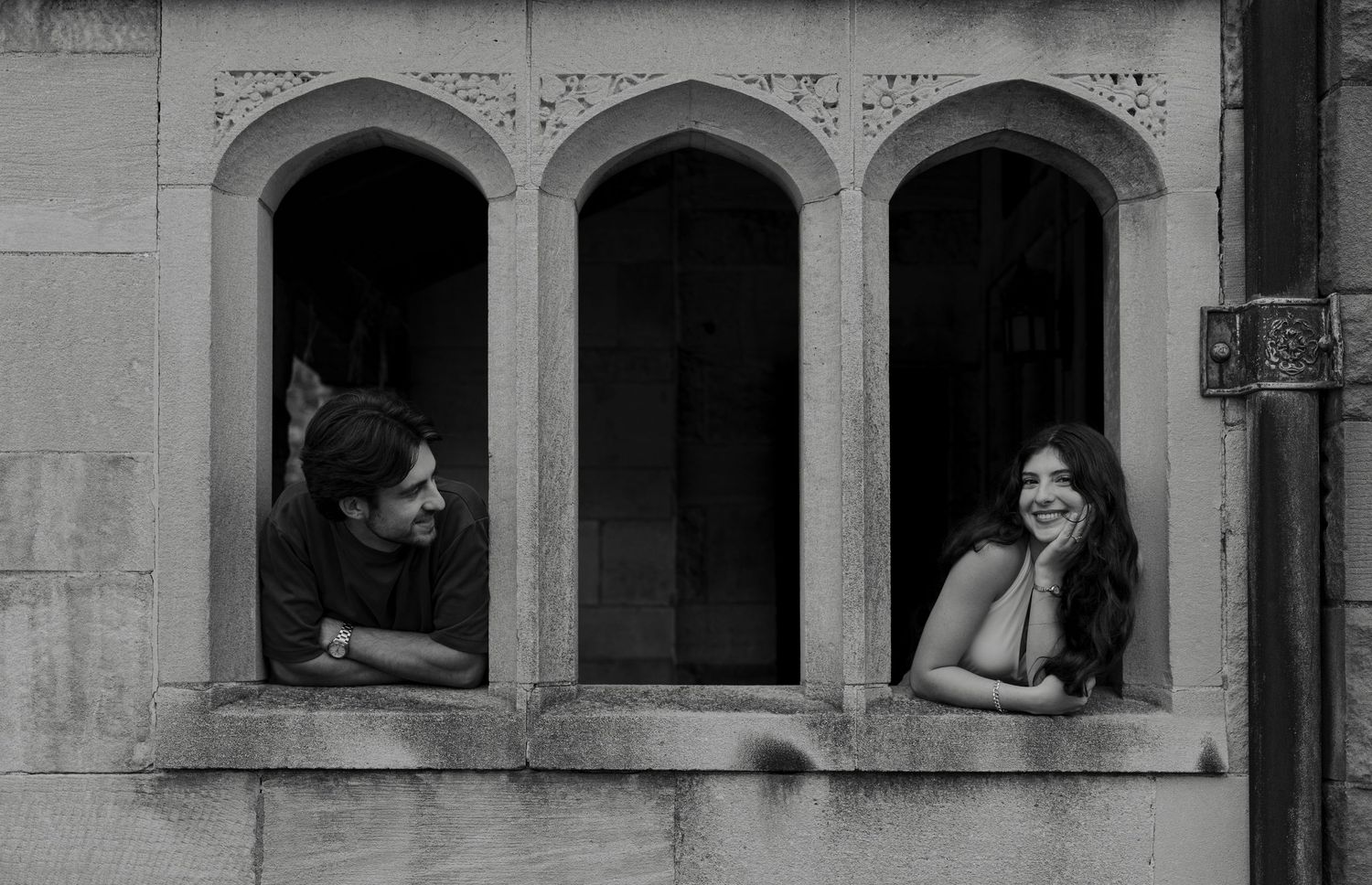 Black and white photo of smiling couple leaning out of arched stone windows together.